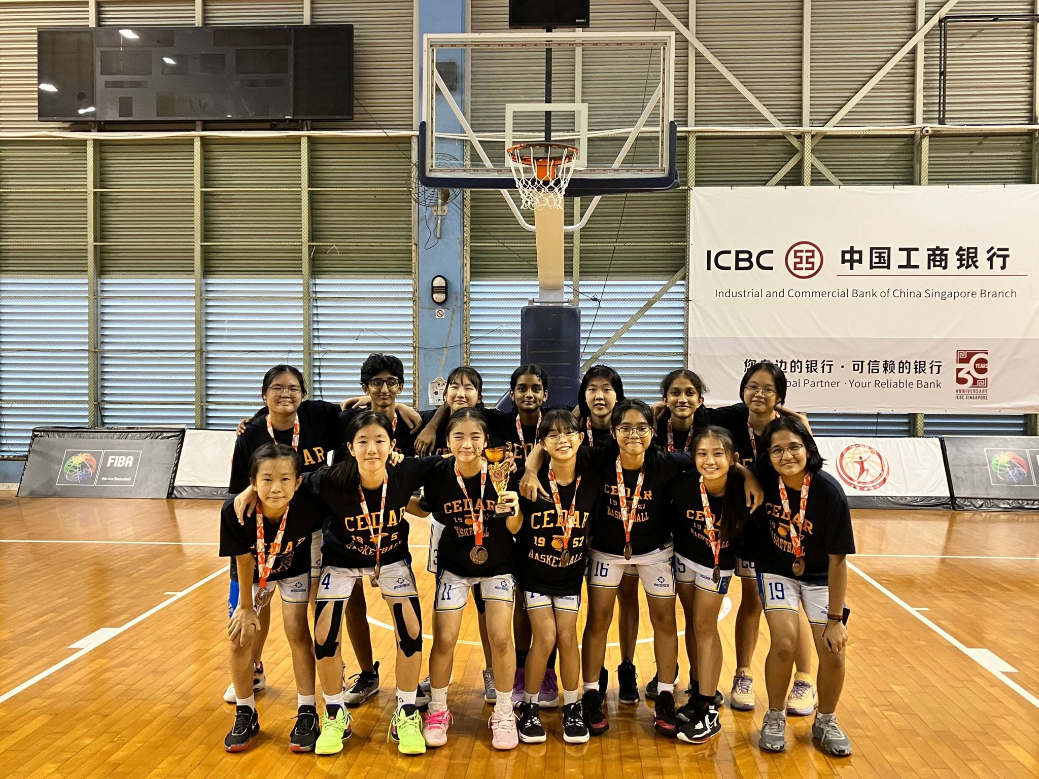 A youth girls basketball team in black Cedar jerseys poses with medals and a trophy on an indoor court.