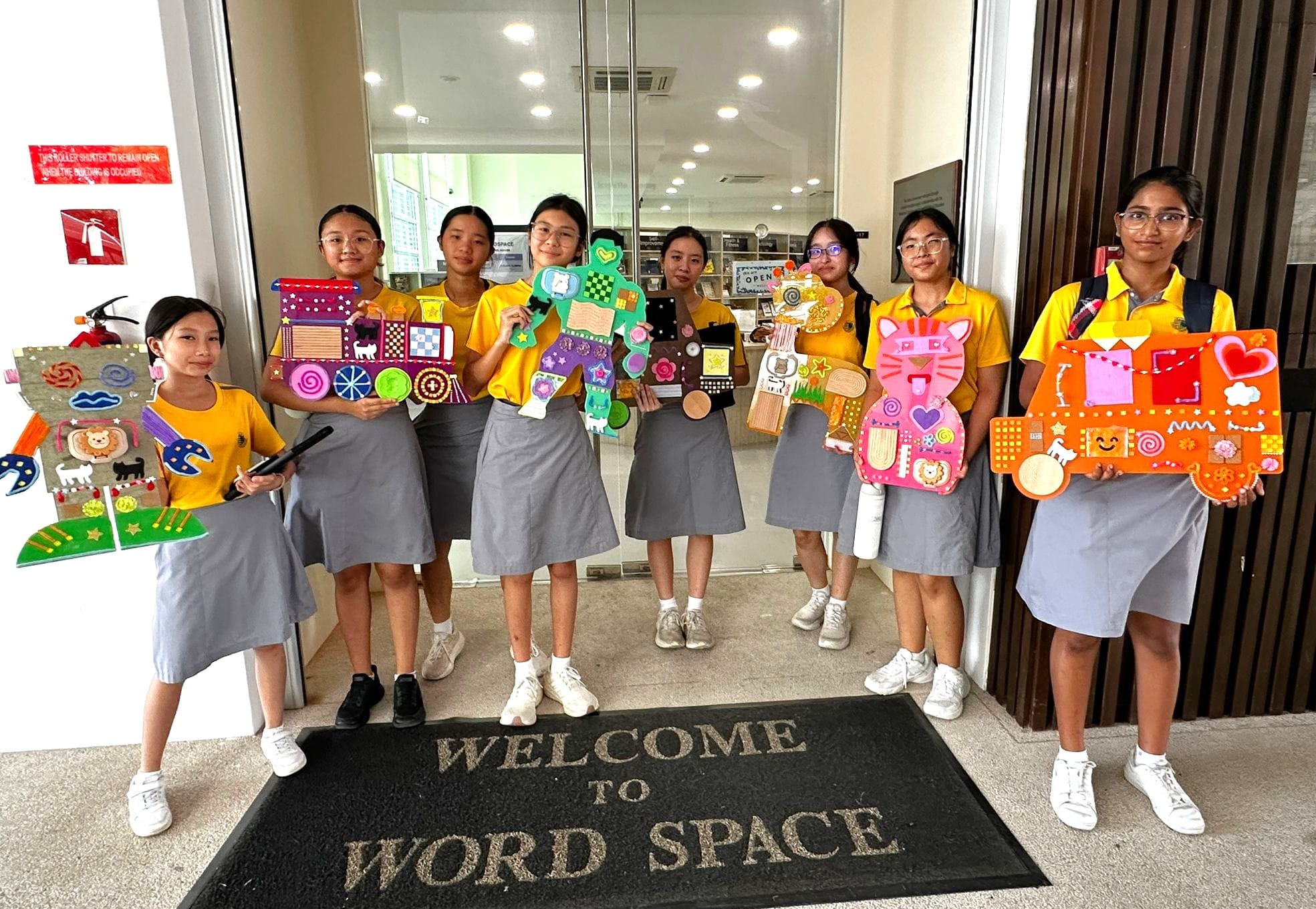 Eight girls in yellow uniforms hold colorful craft projects outside a building with a Welcome to Word Space mat.