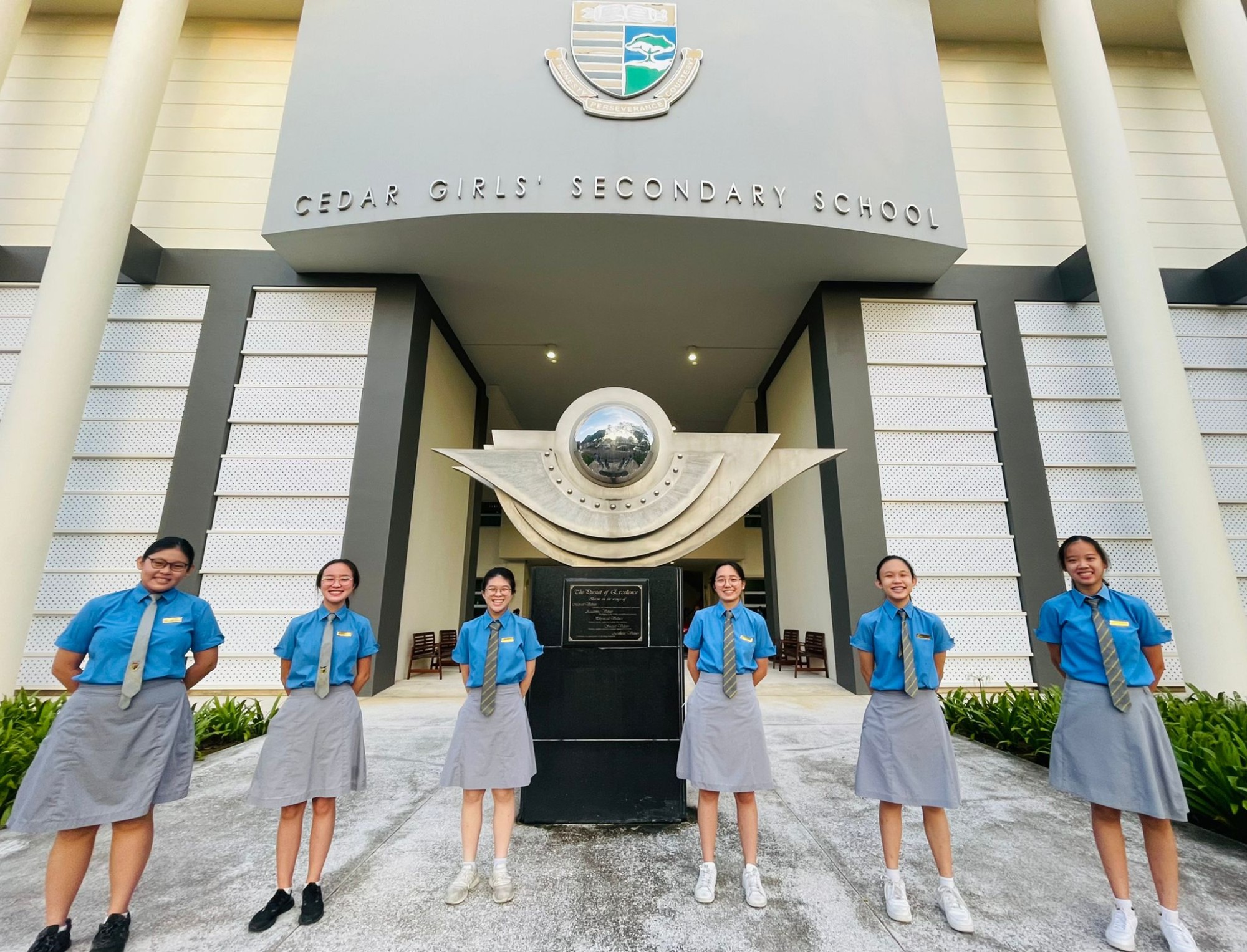 Six girls in blue uniforms stand in front of Cedar Girls' Secondary School entrance with a decorative sculpture.