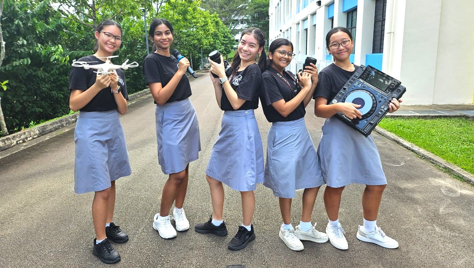 Five schoolgirls in uniforms hold a drone, microphone, camera lens, phone, and DJ controller outdoors.