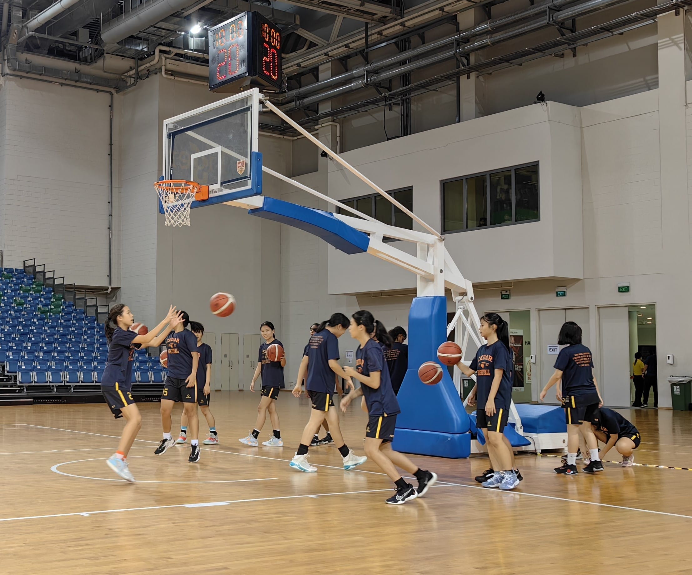 Girls in matching navy uniforms practice basketball drills on an indoor court near a hoop with a scoreboard above.