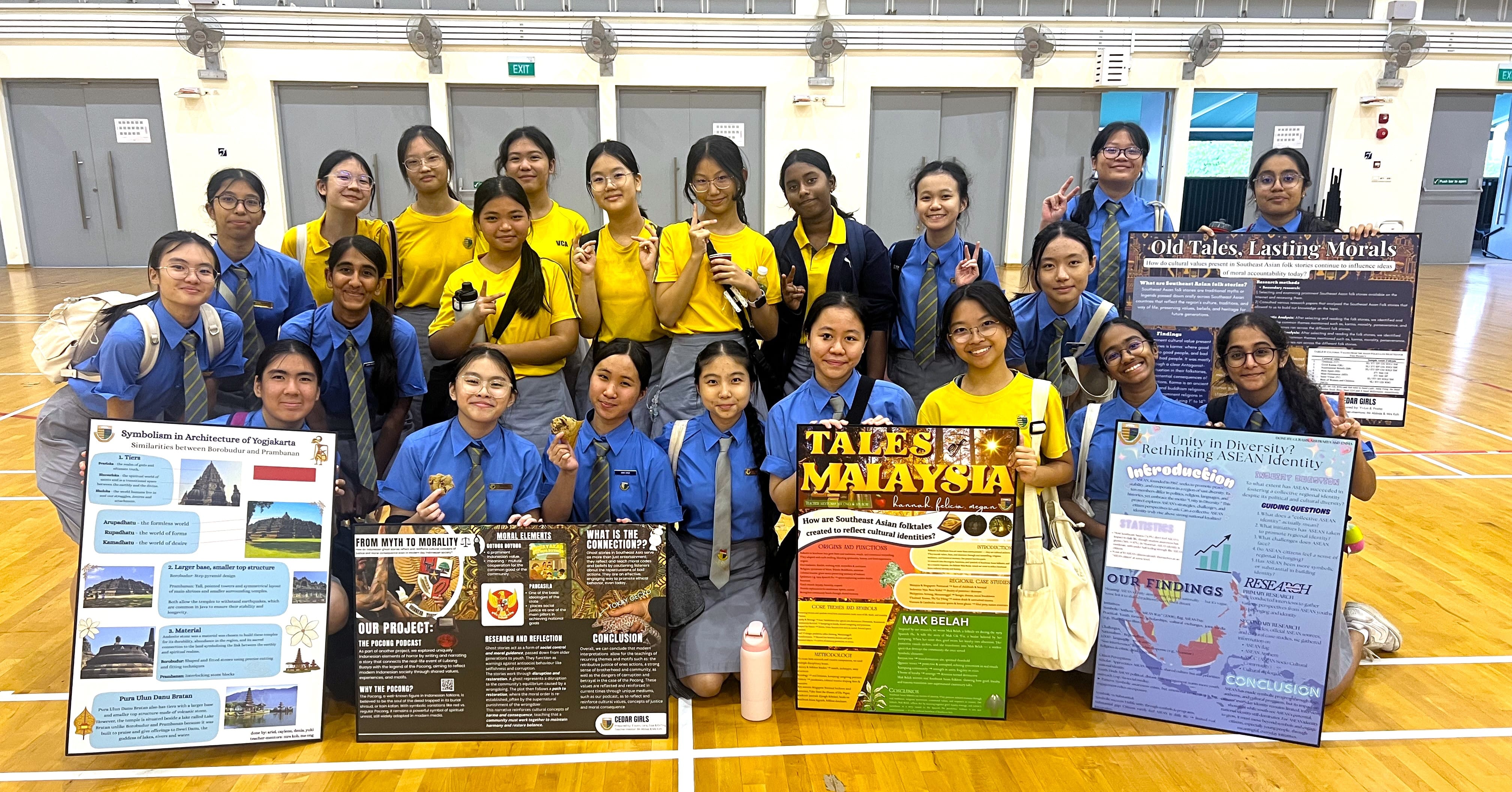 A large group of school girls in blue and yellow uniforms pose holding research project display boards in a gym.