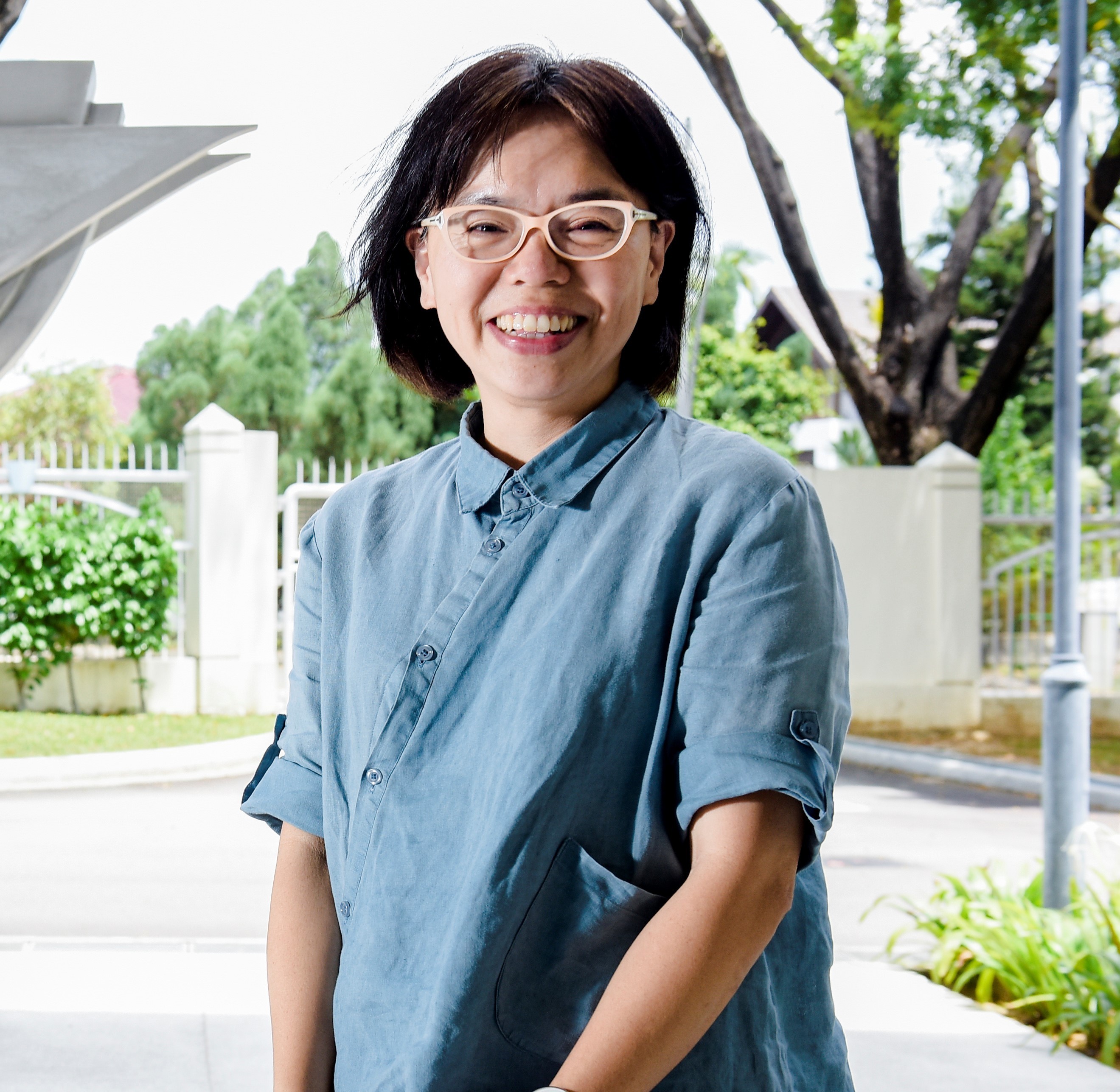 A smiling woman with short black hair and cream glasses wearing a blue linen shirt stands outdoors near trees.