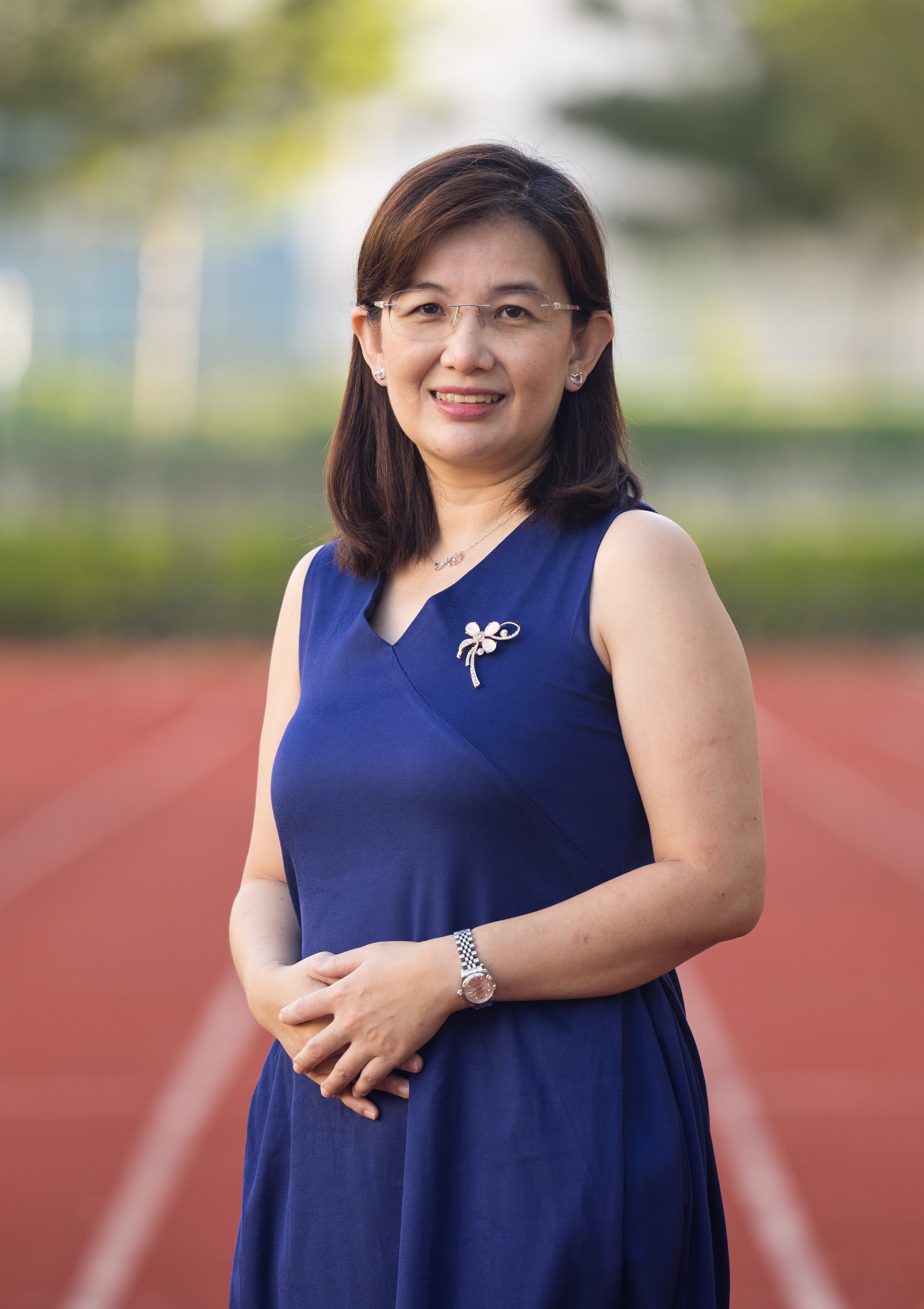 A smiling woman in a blue sleeveless dress with a brooch and glasses stands on a red running track.