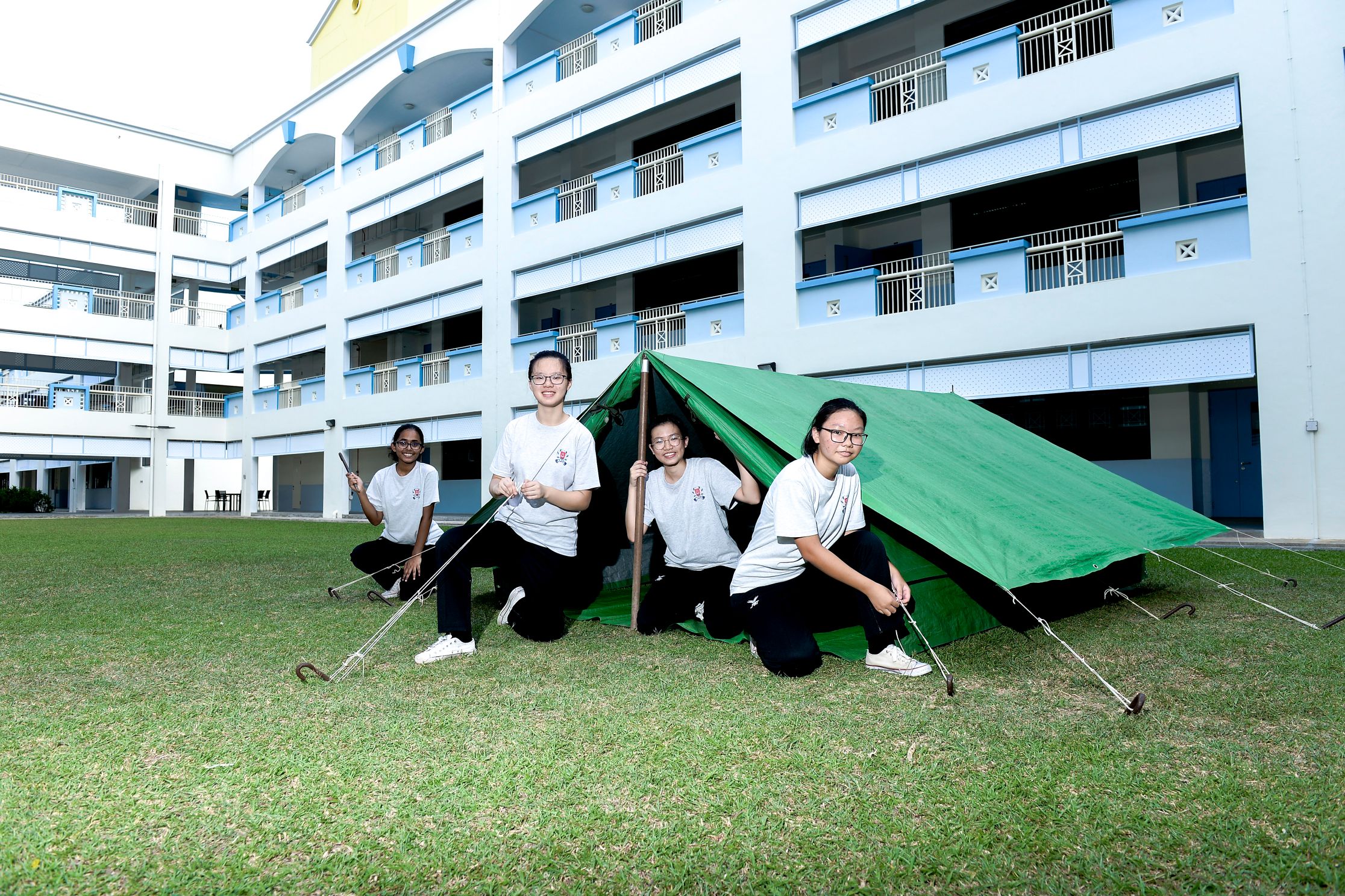 Four students in white shirts pose with a green tent pitched on grass in a school courtyard.