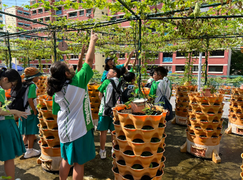 Children in green uniforms explore an outdoor garden with stacked terracotta planters and a vine-covered trellis overhead.