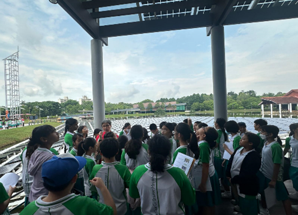 Students in green uniforms gathered under an outdoor pavilion near a large solar panel field on a cloudy day.
