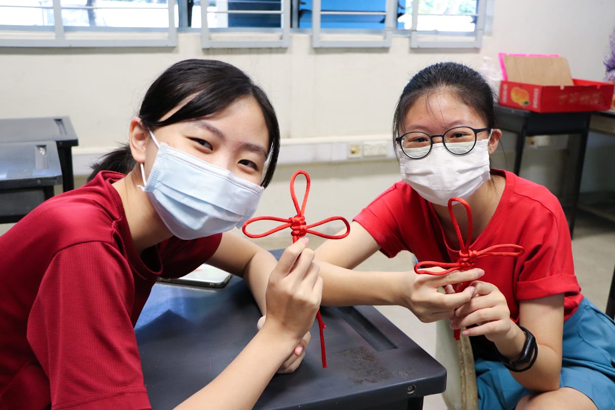 Two masked girls in red shirts hold up handmade red knotted cord decorations in a classroom.