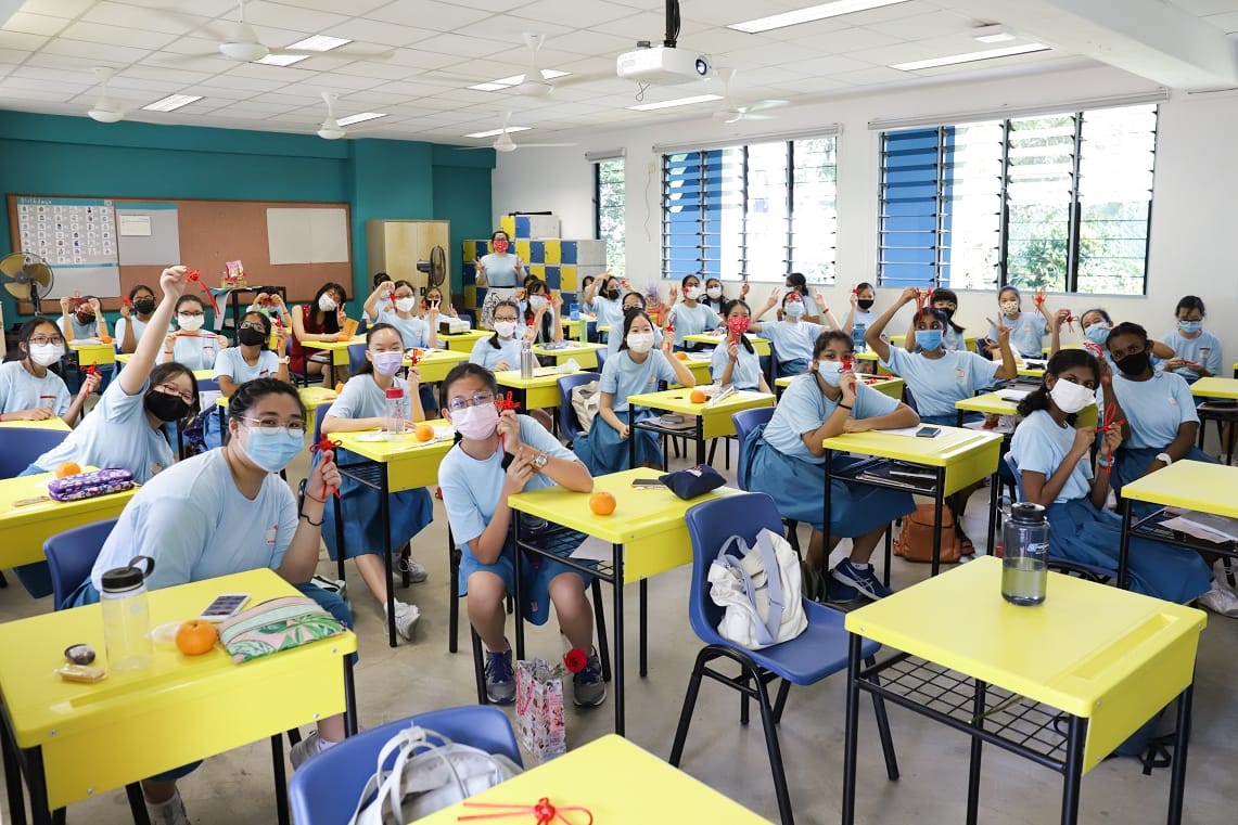 Students in light blue uniforms and face masks sit at yellow desks, holding up red decorative items in a classroom.