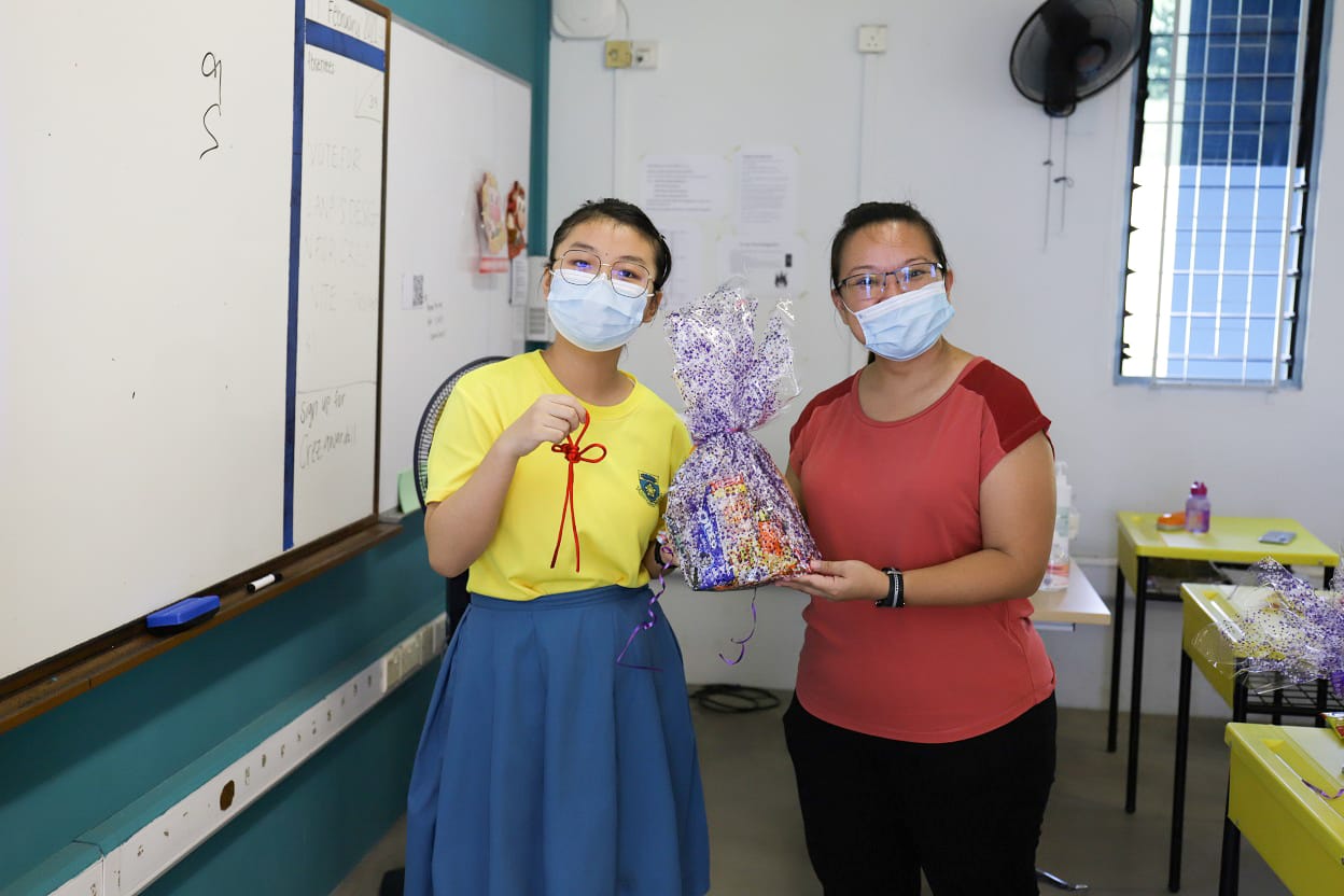 Two masked girls stand in a classroom holding a purple wrapped gift bag near a whiteboard.