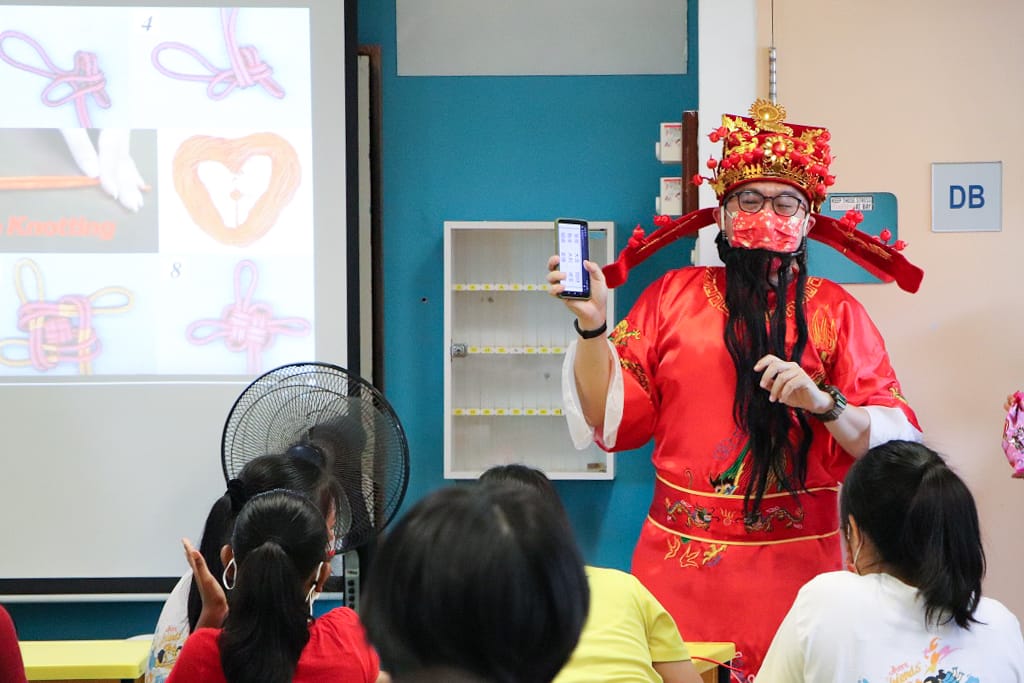 Person in red Chinese traditional costume and crown holds a phone while presenting a knotting slideshow to seated students.