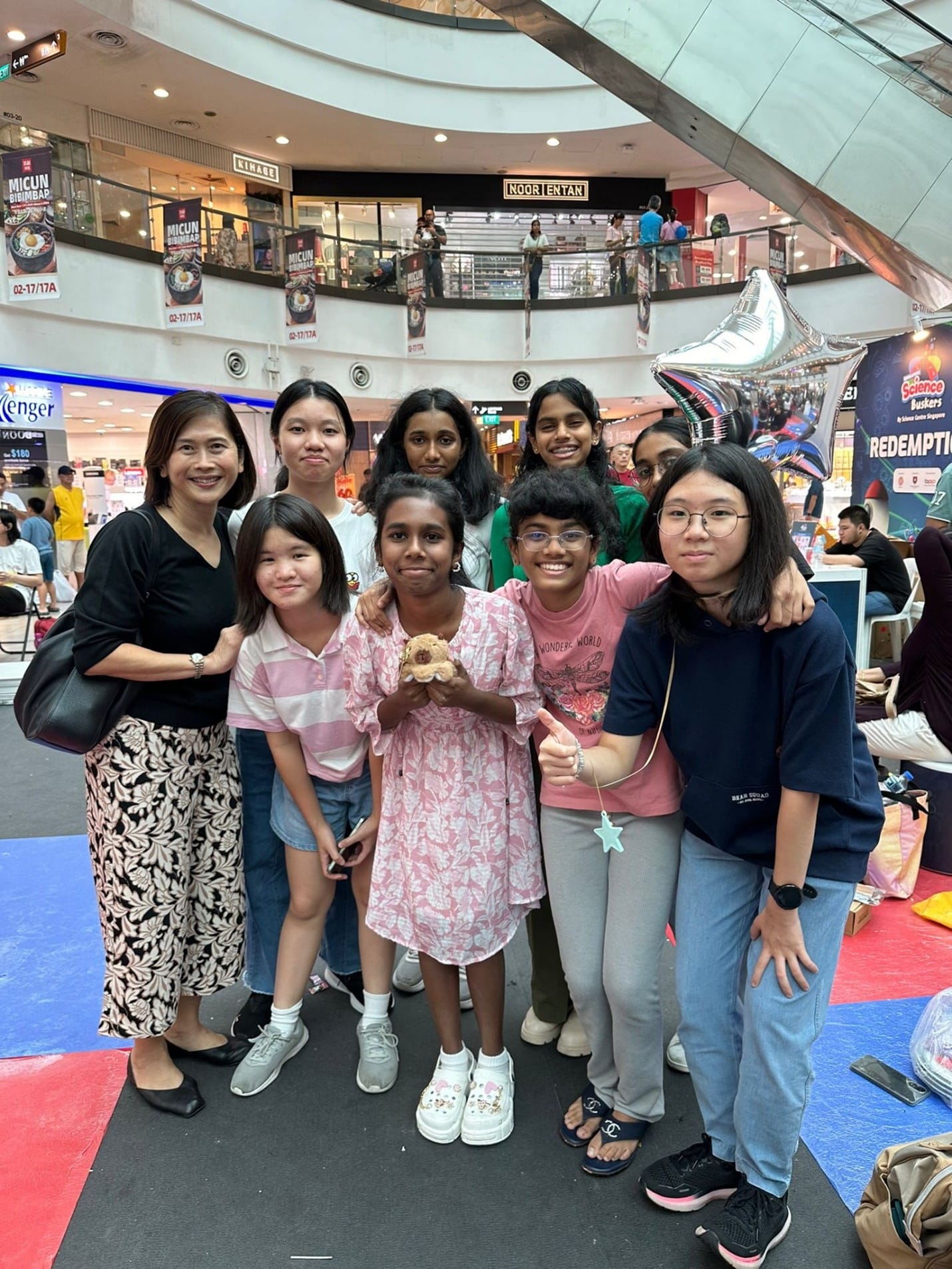 A group of nine people, including children and adults, posing together inside a busy shopping mall.