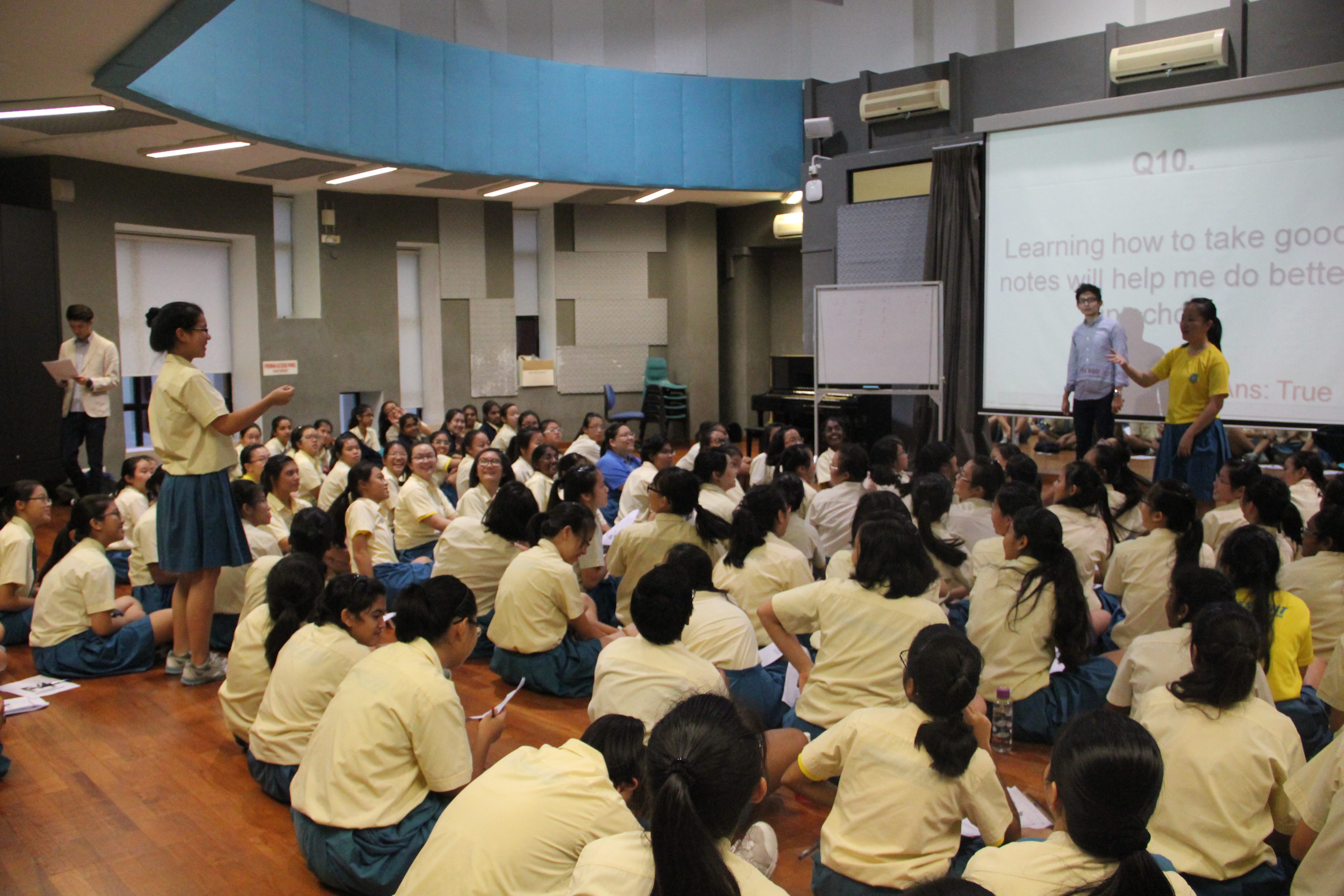 Large group of students seated on floor facing a projector screen while two presenters speak at front.