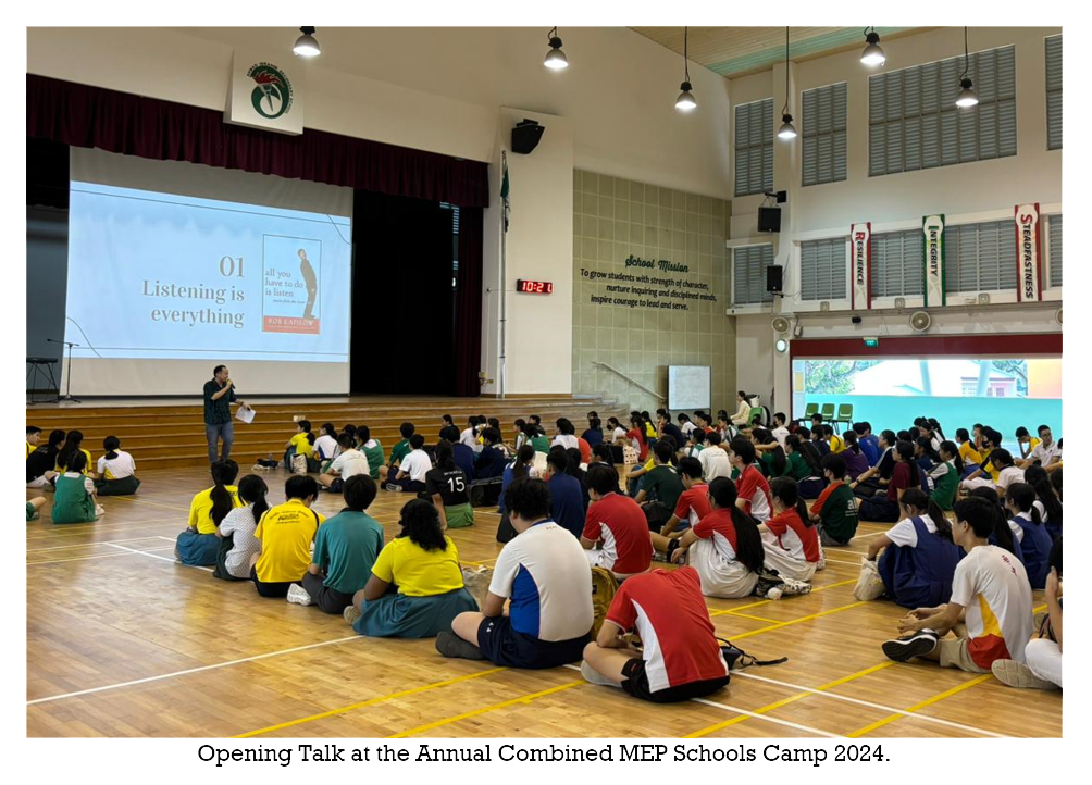 Students sit on a gymnasium floor facing a speaker and a screen reading "Listening is everything."