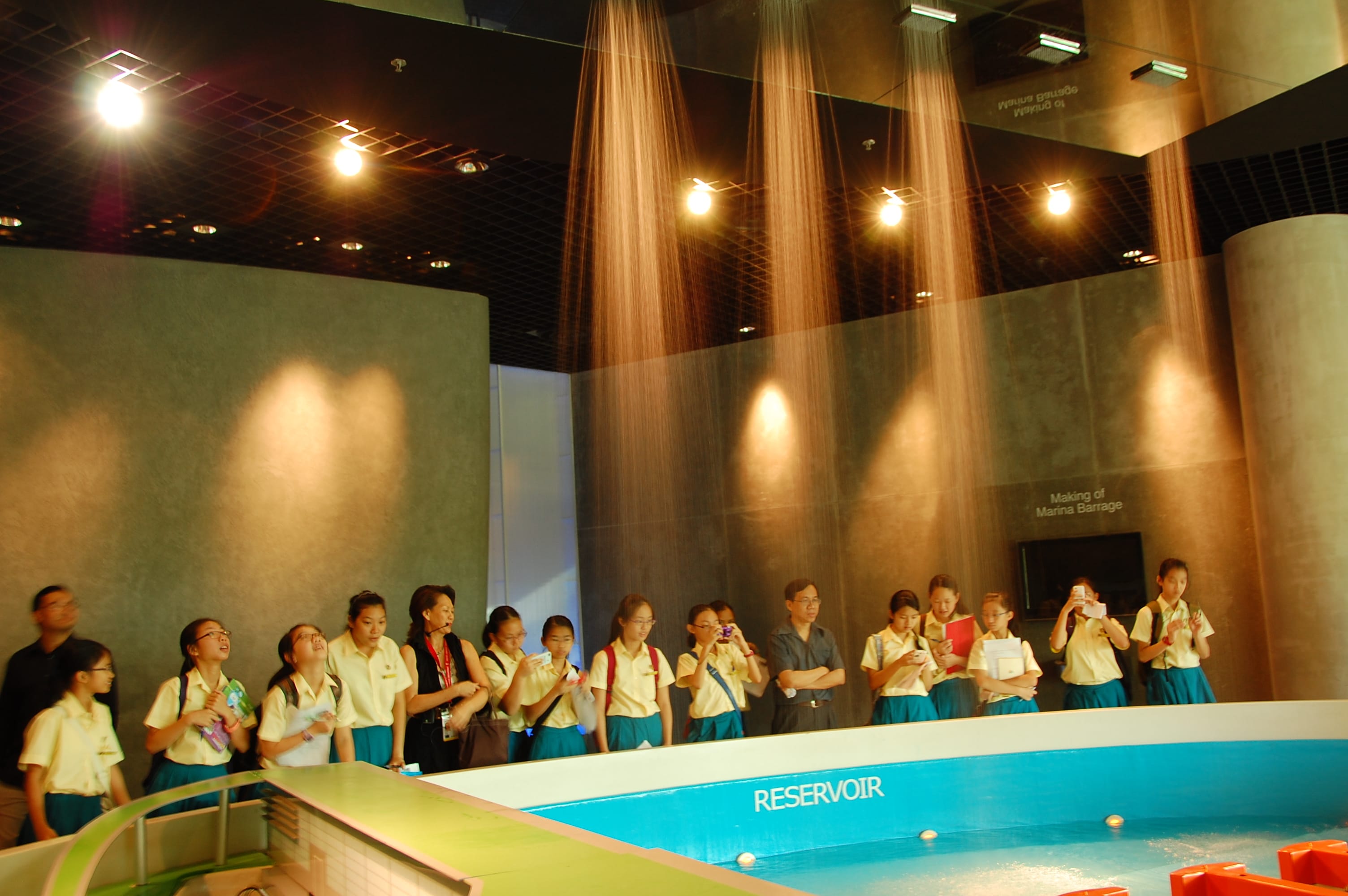 Students in uniform stand around a reservoir exhibit at the Marina Barrage museum with indoor rain curtains above.