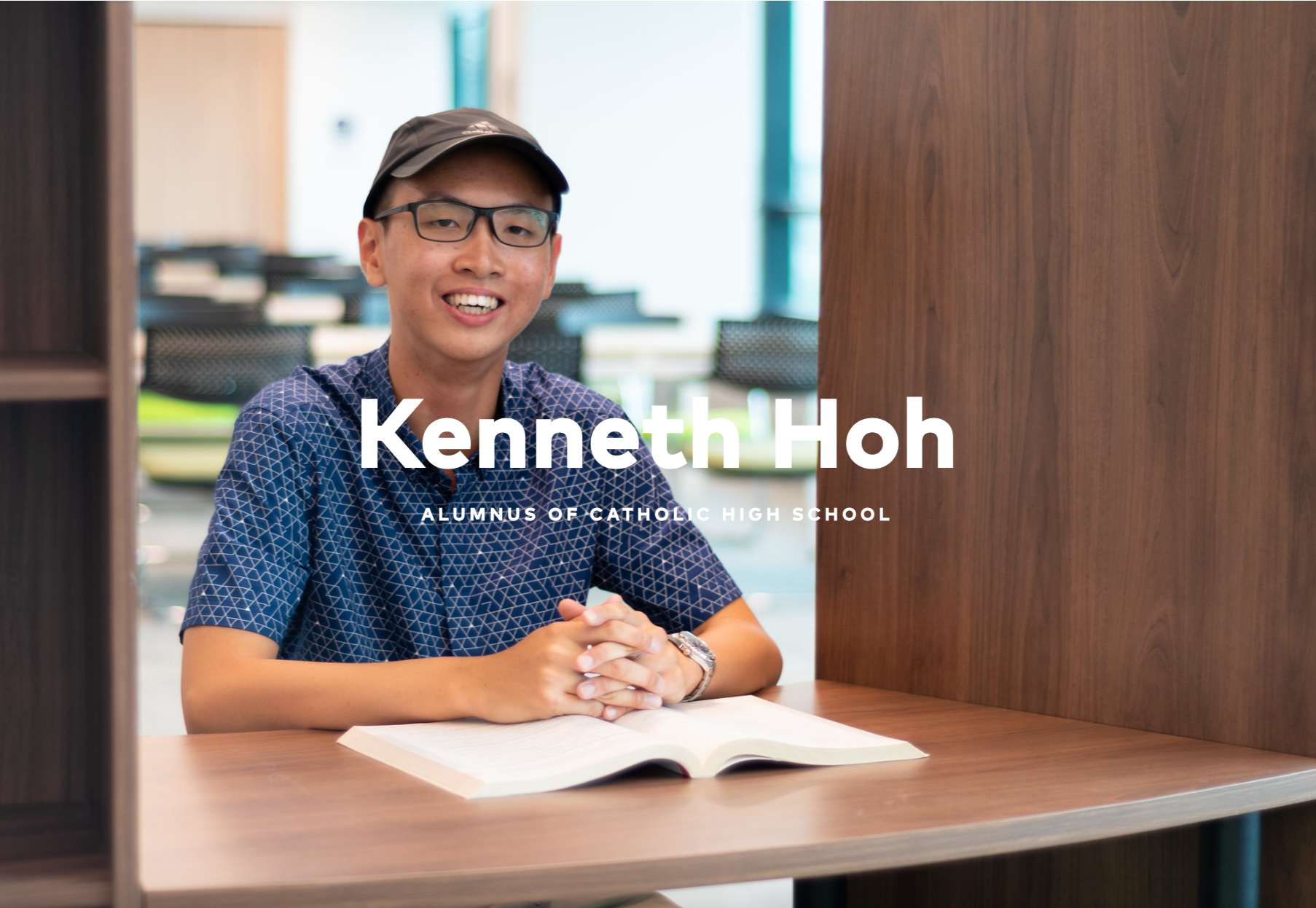Smiling young man with glasses and a cap sitting at a desk with an open book, labeled Kenneth Hoh, Catholic High School alumnus.