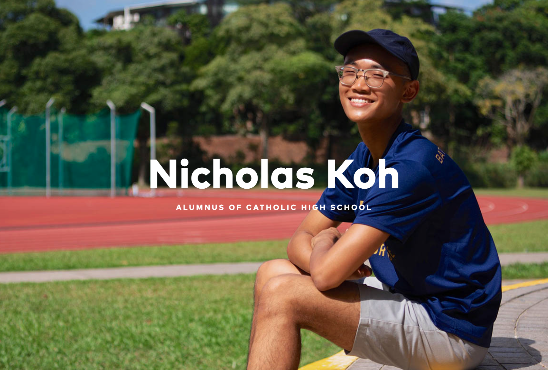 Nicholas Koh, a smiling young man with glasses and a navy cap, sits outdoors near a red running track.