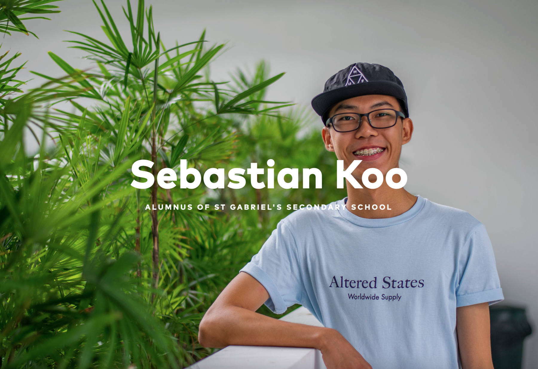 Young man named Sebastian Koo smiling, wearing glasses, a black cap, and a light blue shirt, standing near green plants.