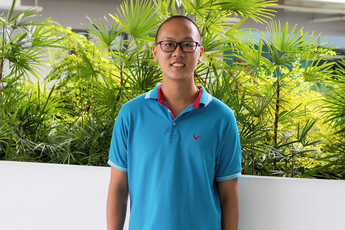 Young man with glasses and short hair wearing a teal polo shirt, smiling in front of lush green tropical plants.