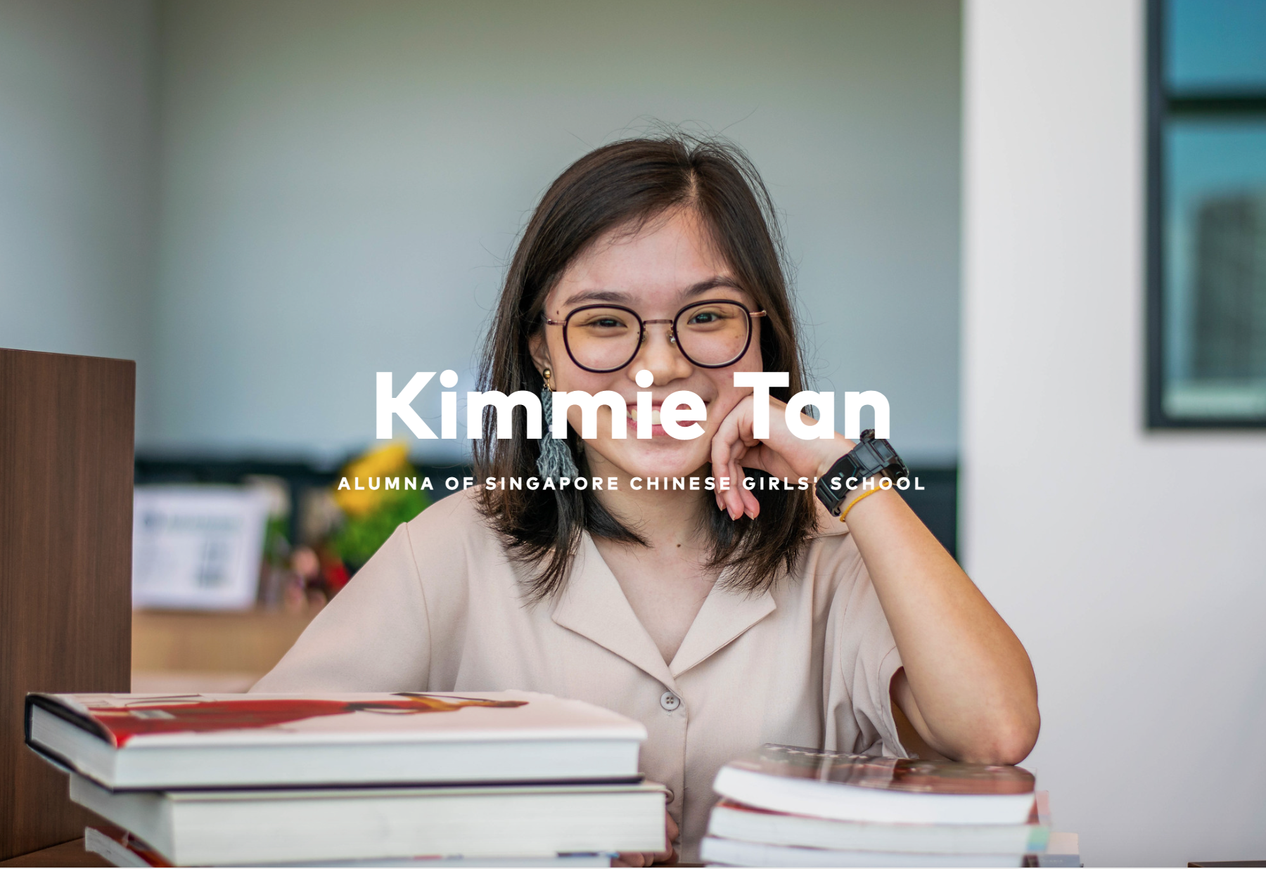 Smiling young woman with glasses seated at a desk with stacked books, labeled Kimmie Tan, alumna of Singapore Chinese Girls' School.