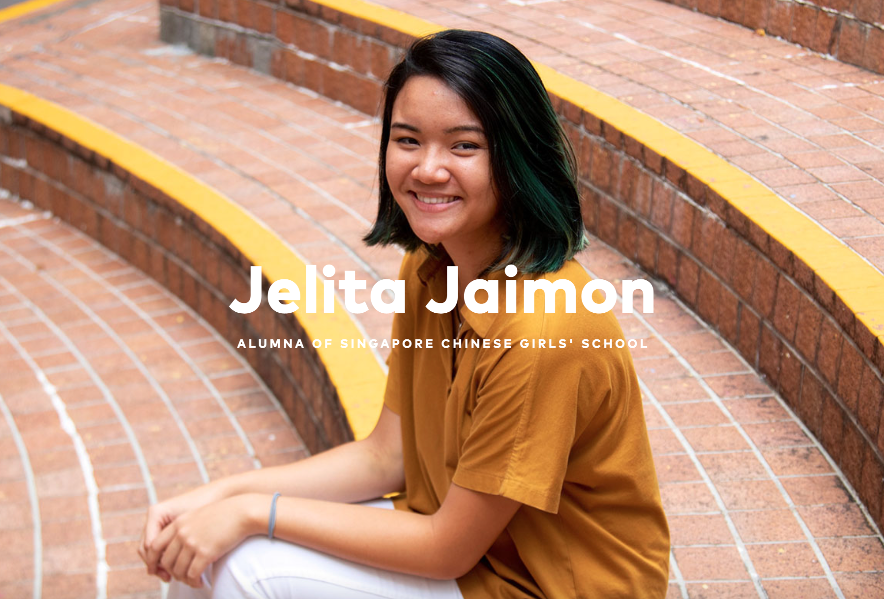 Jelita Jaimon, a smiling young woman with dark teal-tipped hair, sits on brick steps wearing a mustard yellow shirt.
