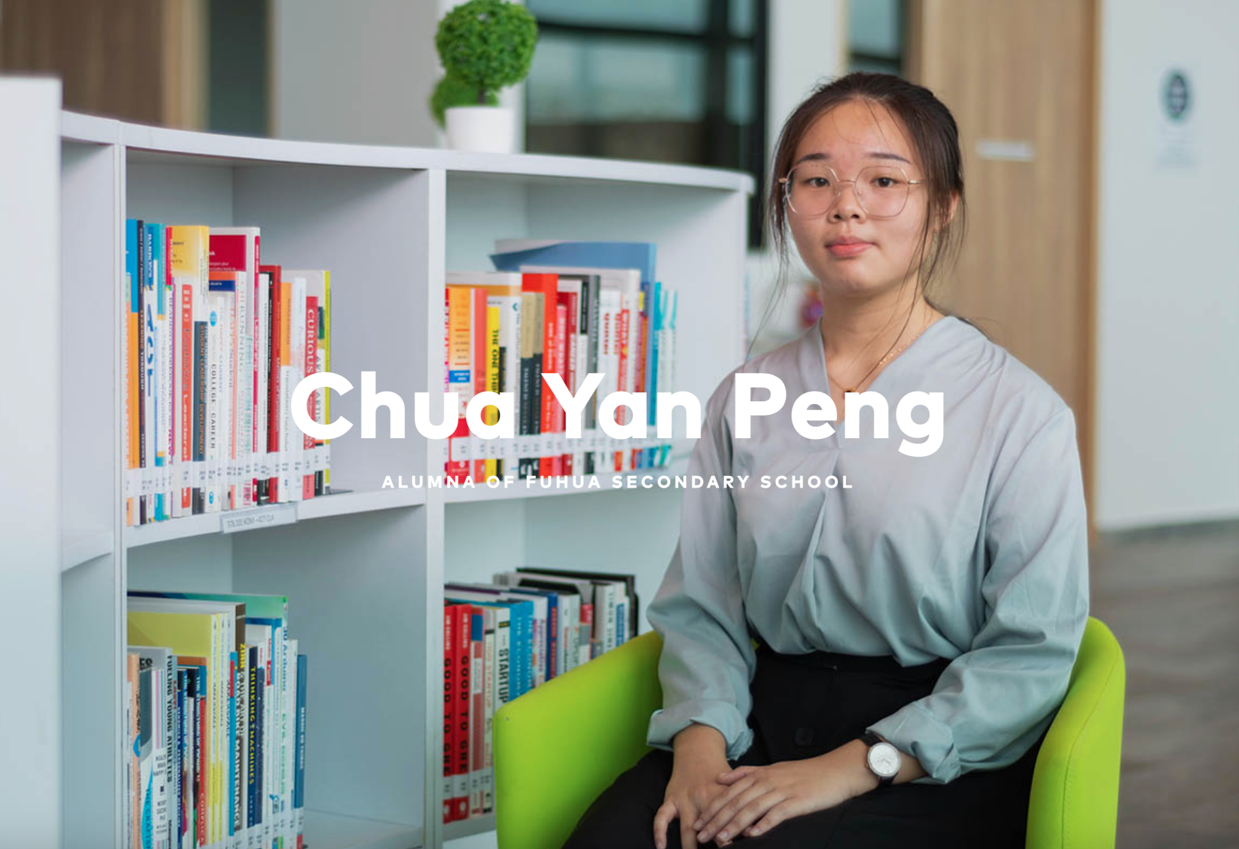 Young woman with glasses sits in a green chair beside a white bookshelf filled with colorful books.