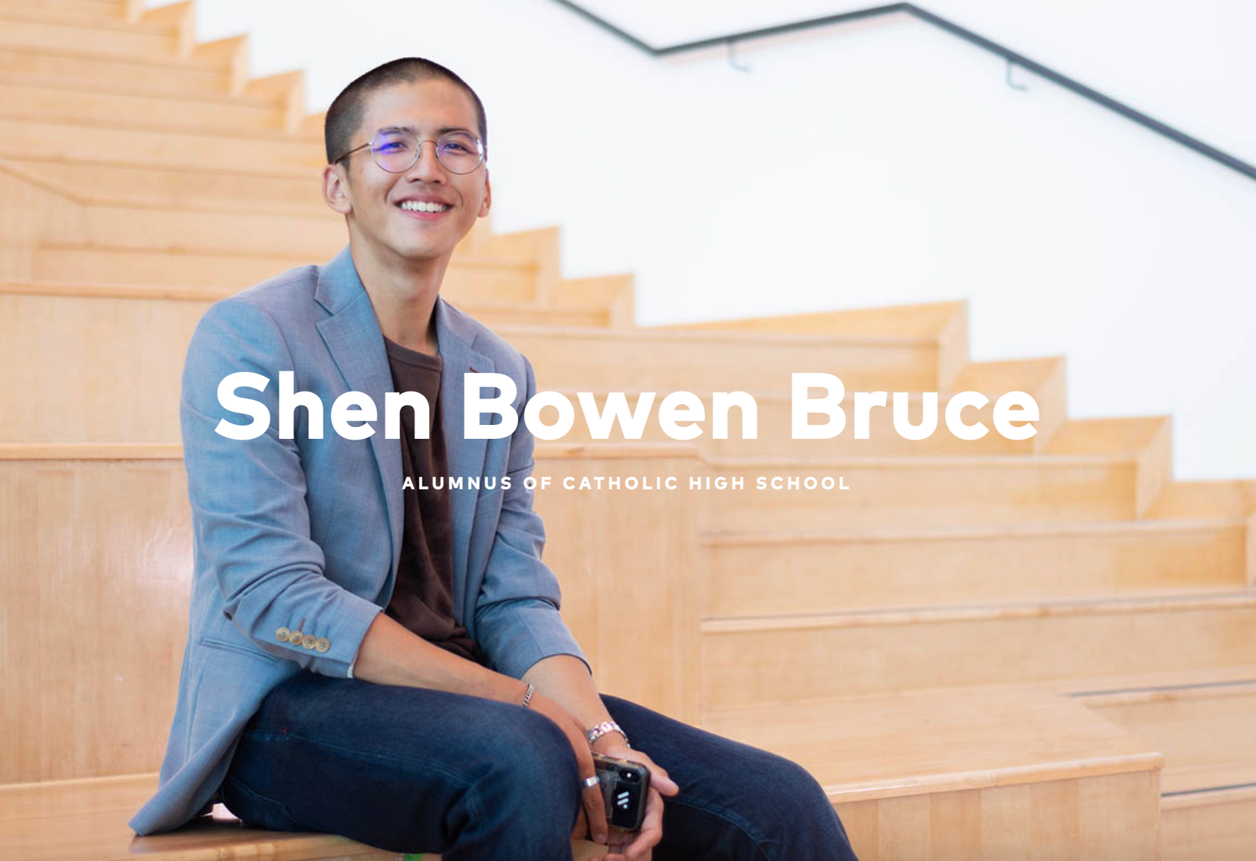 Shen Bowen Bruce, a smiling young man with glasses and a blazer, sits on wooden stairs.