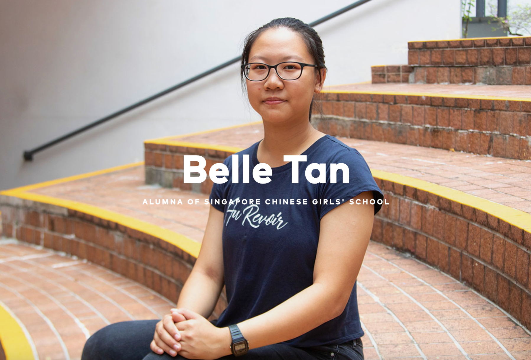 Young woman with glasses in a navy shirt sits on brick steps, labeled Belle Tan, alumna of Singapore Chinese Girls' School.