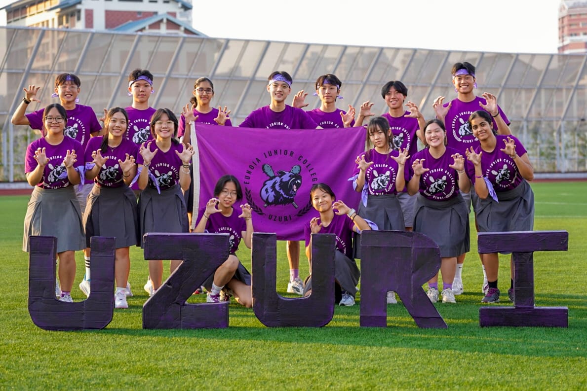 Students in purple Eunoia Junior College shirts pose on a field with a flag and large letters spelling UZURI.