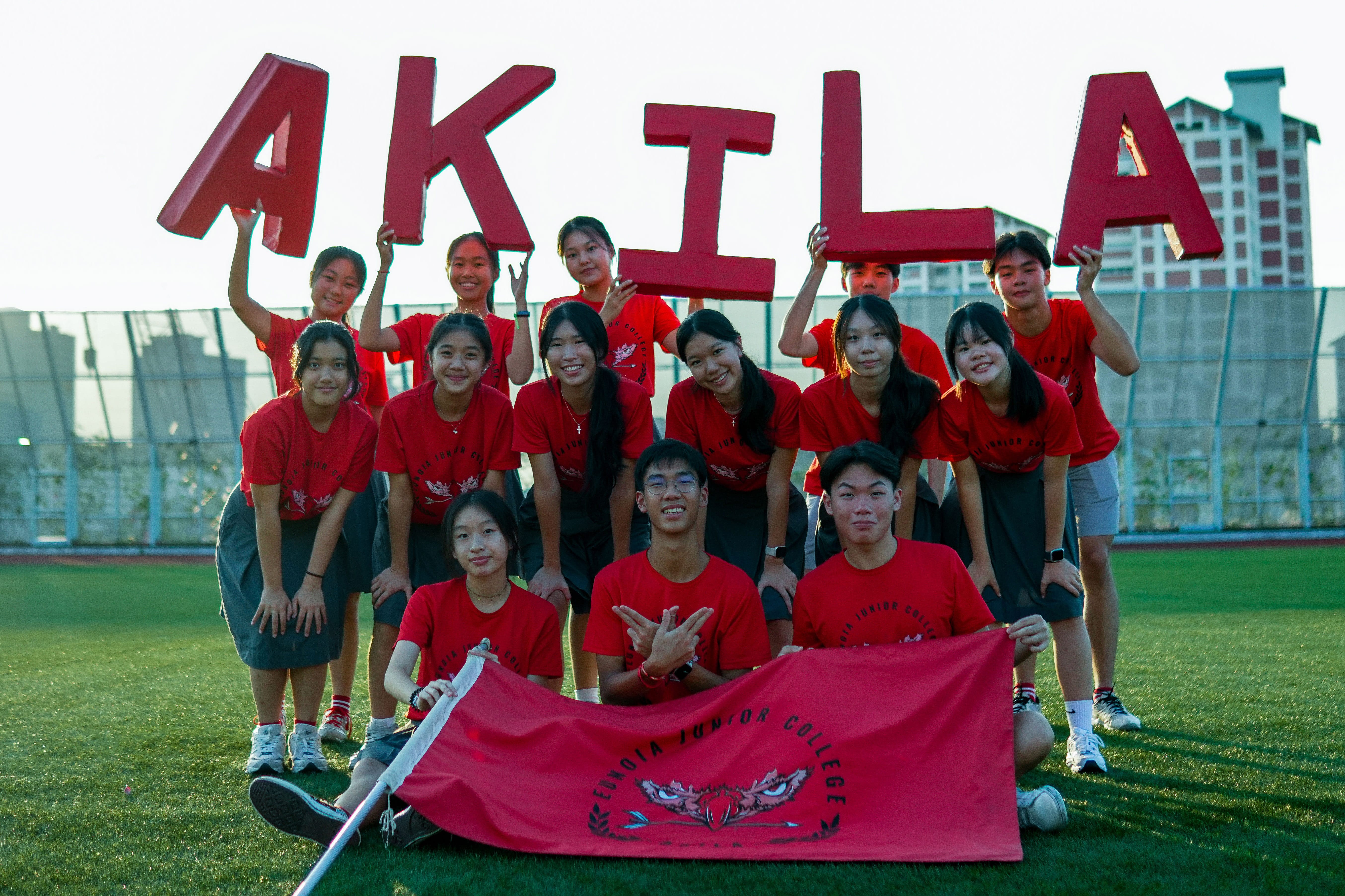 Group in red shirts on a field holding large letters spelling AKILA, with a red banner in front