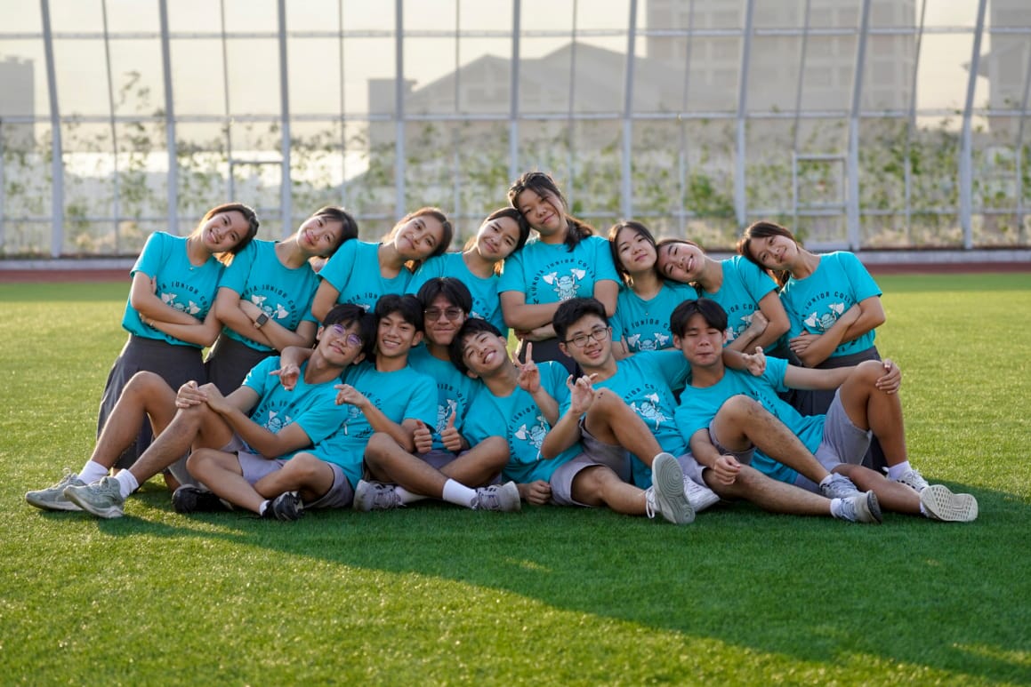 A group of about 14 young people in matching teal shirts posing together on a green field, smiling at the camera.