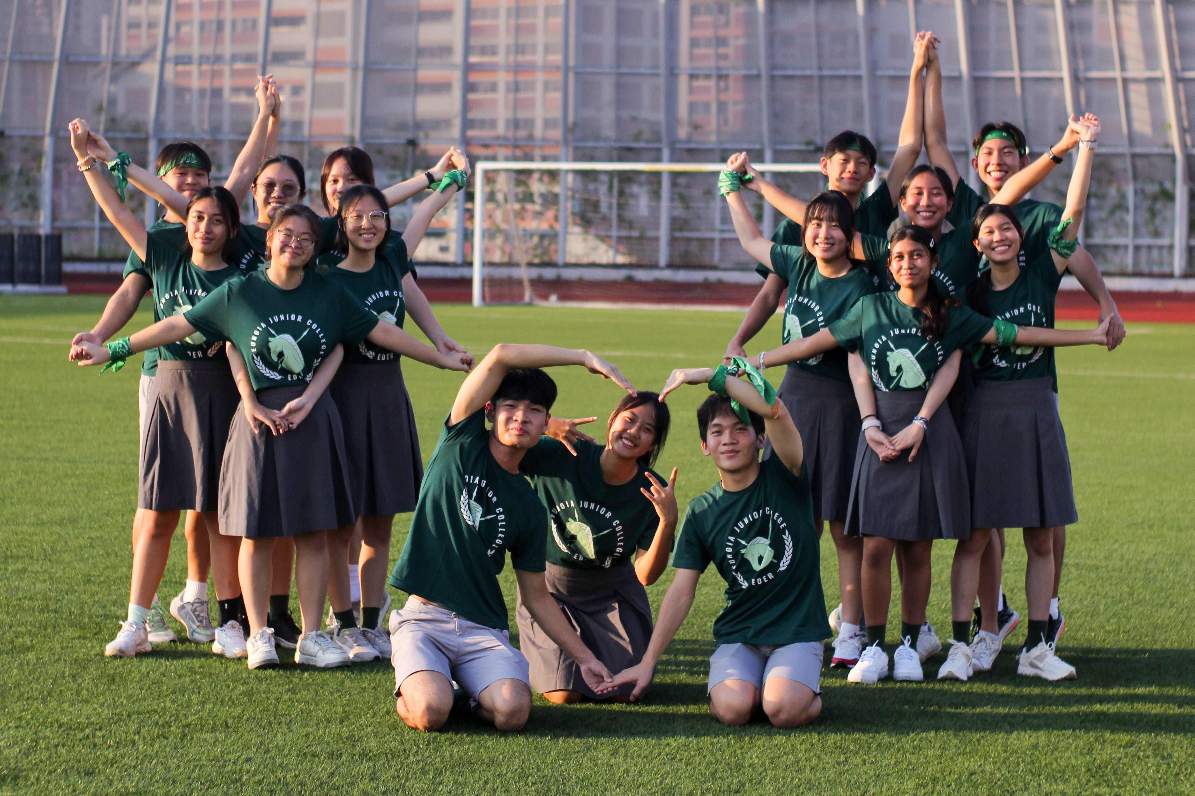 Group of students in green shirts and gray skirts pose on a sports field with arms raised in patterns