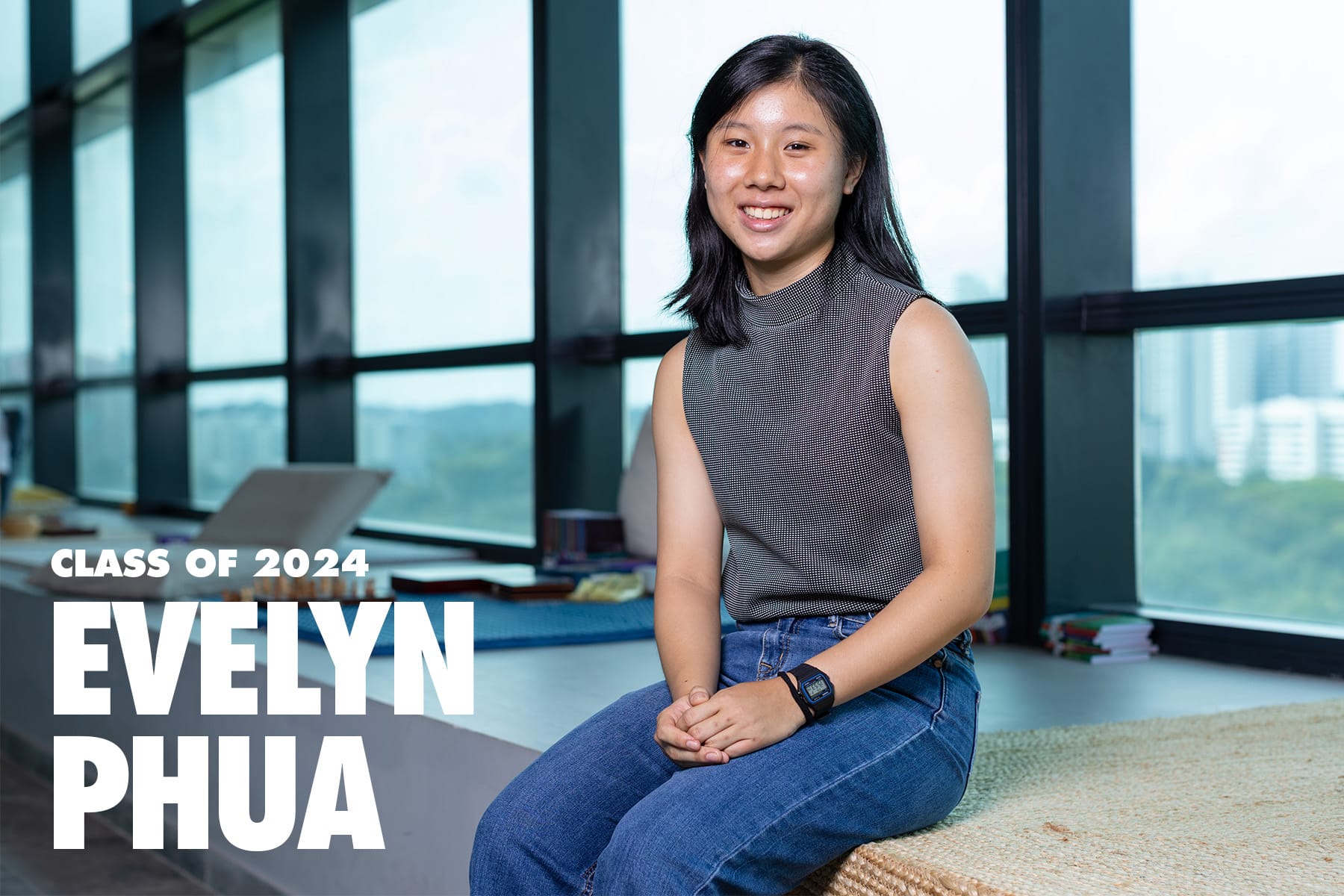 Evelyn Phua, Class of 2024, smiling young woman in a grey top and jeans seated near large windows.