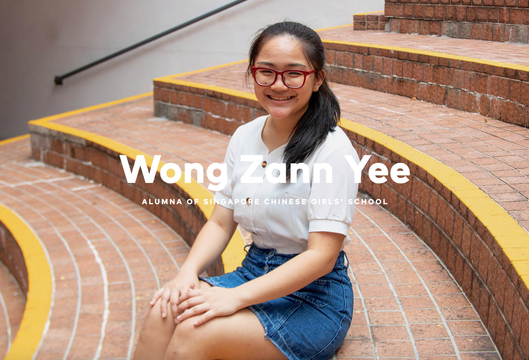 Young woman with red glasses and dark hair sits smiling on brick steps with yellow edges, wearing a white top and denim skirt.