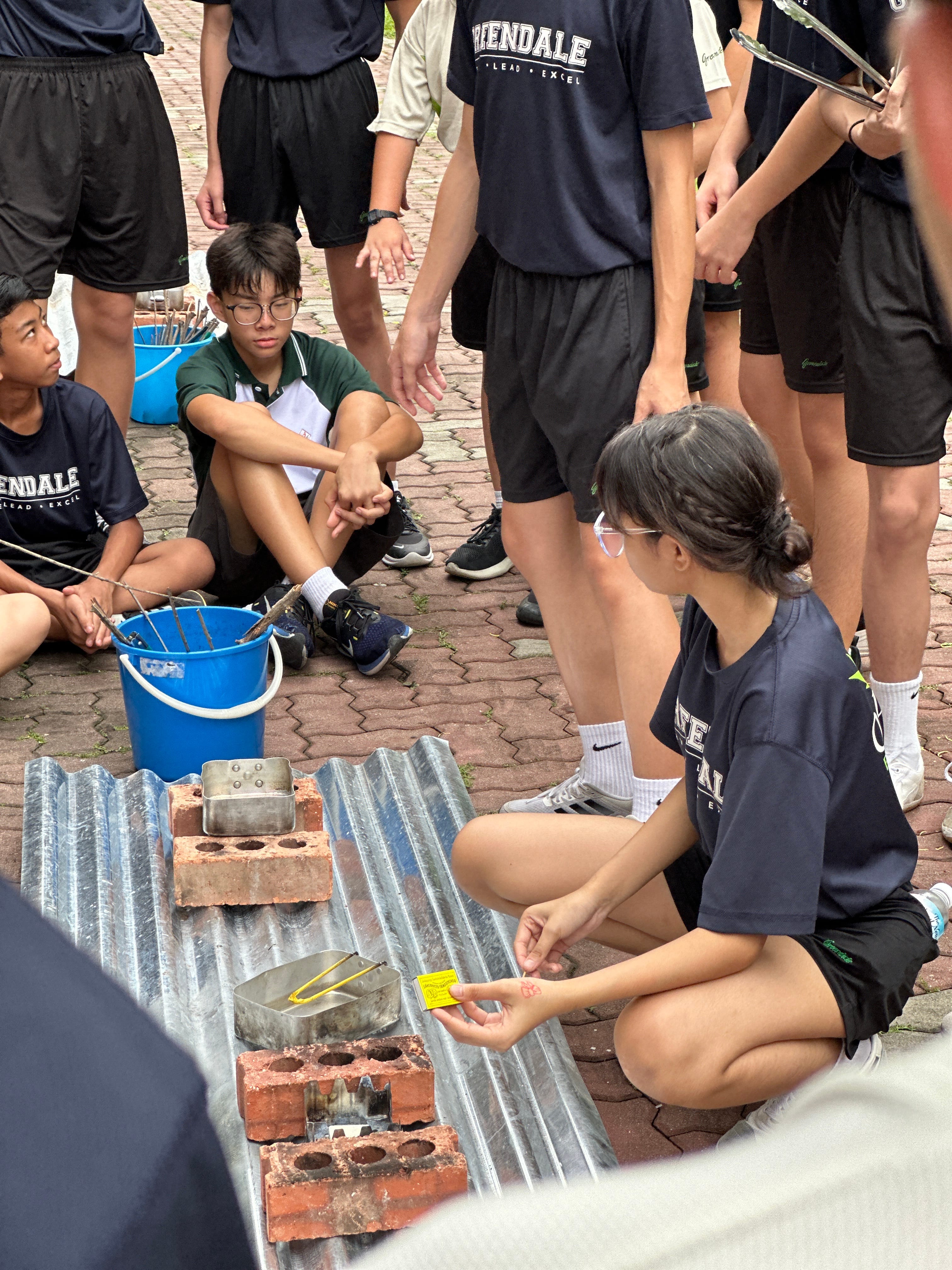 A female student at outdoors, using matches to light an outdoor cooking setup made of bricks and metal trays, while a group of peers watches.