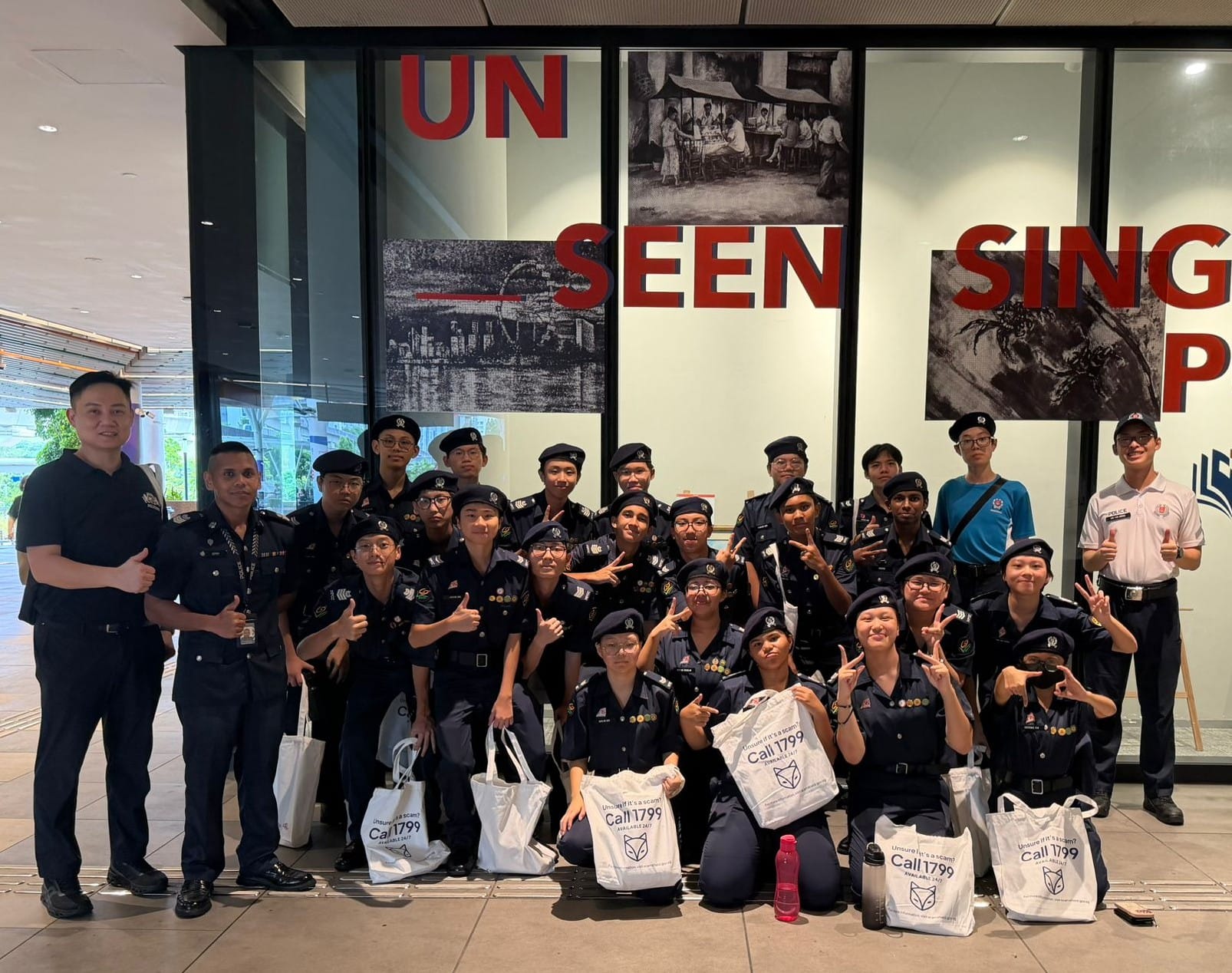 A large group of uniformed officers pose smiling in front of an Unseen Singapore display, holding Call 1799 tote bags.