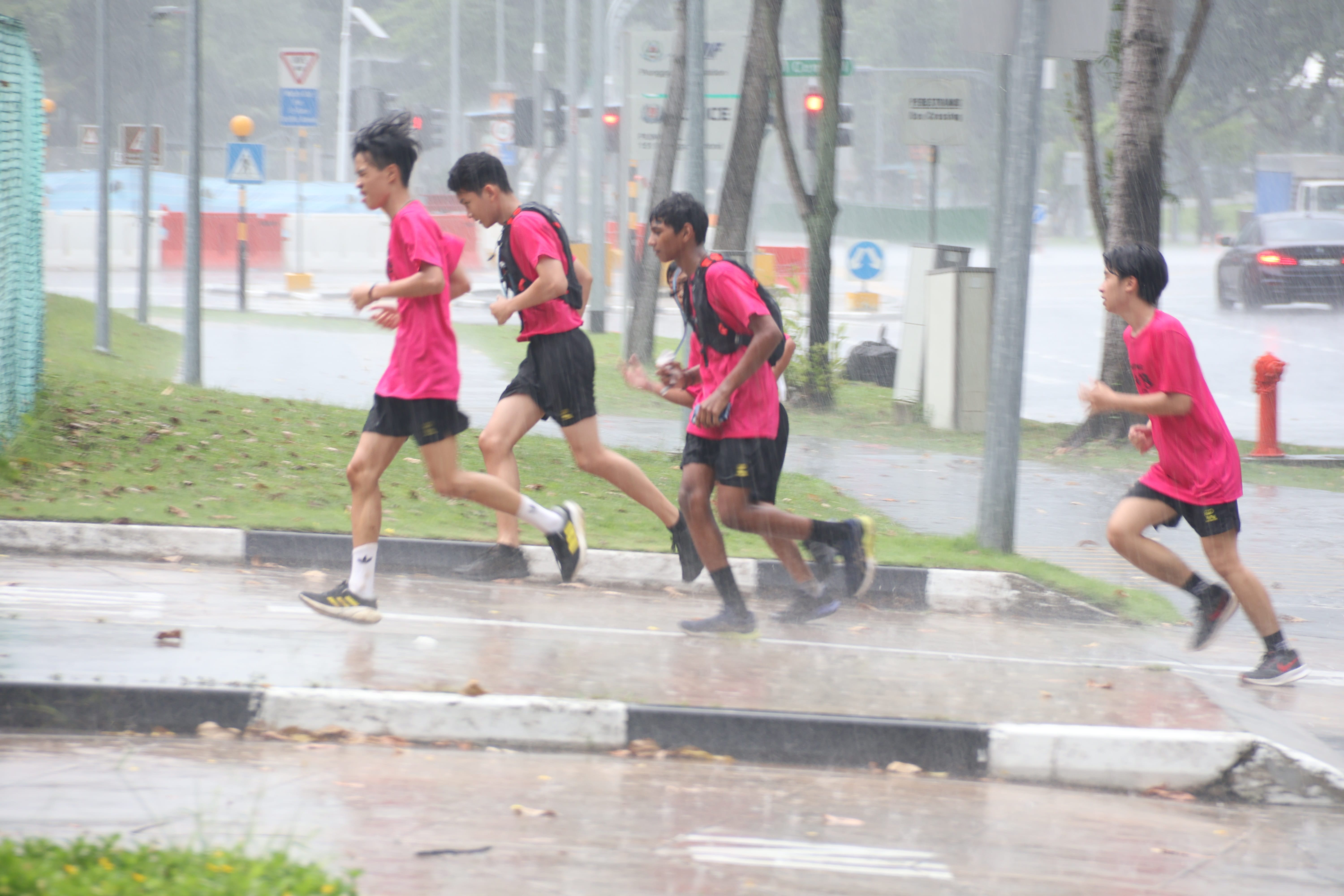A group of students wearing pink t-shirts run together along a wet pavement next to a road during a heavy downpour.