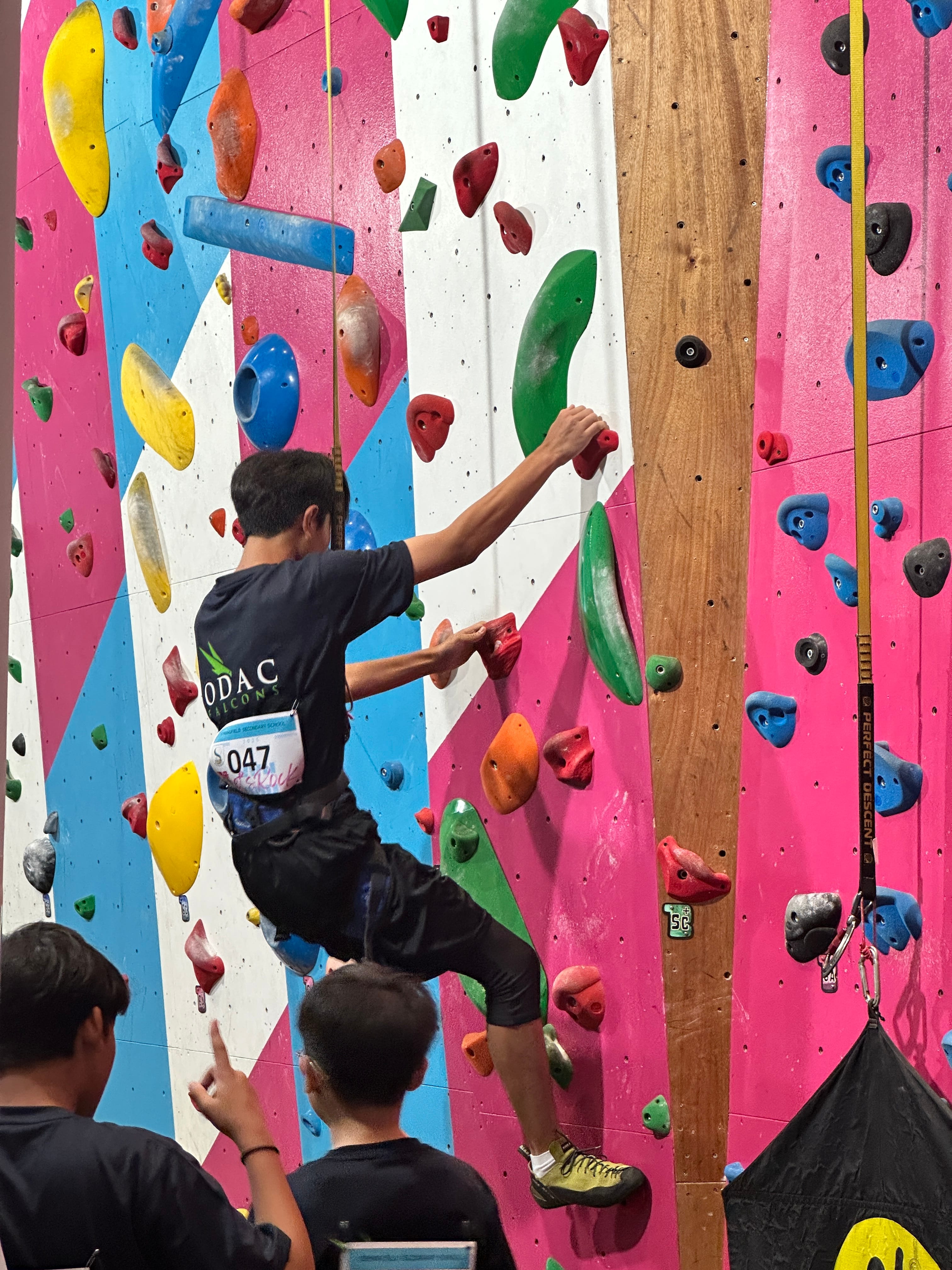 A climber wearing a black "ODAC" shirt ascends a brightly colored, multi-patterned indoor rock climbing wall while onlookers watch from below.