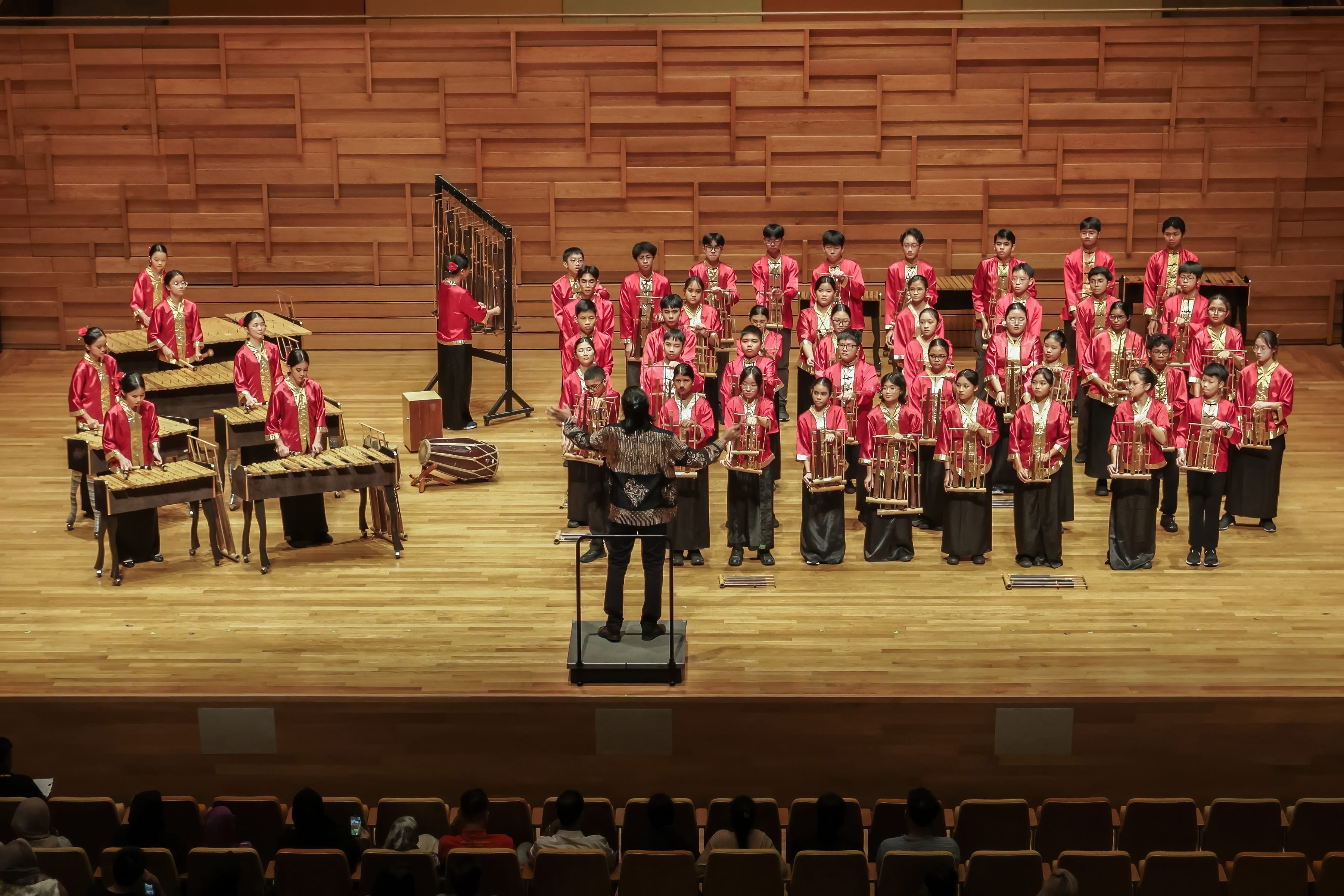 A conductor leads a large youth ensemble in red robes playing traditional instruments on a wooden concert stage.