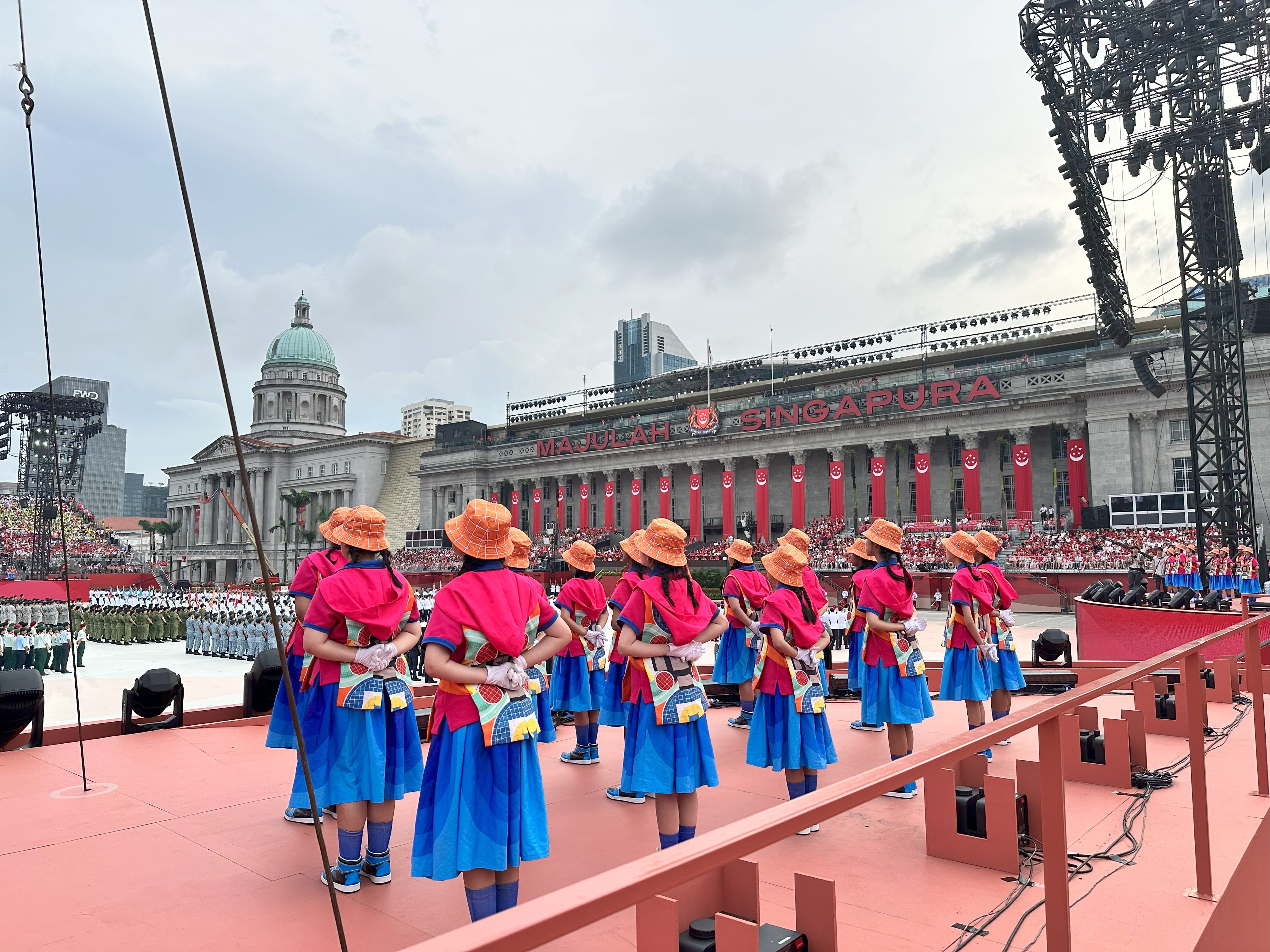 Girls in pink tops, blue skirts, and orange hats stand on a red stage at a Singapore National Day parade.
