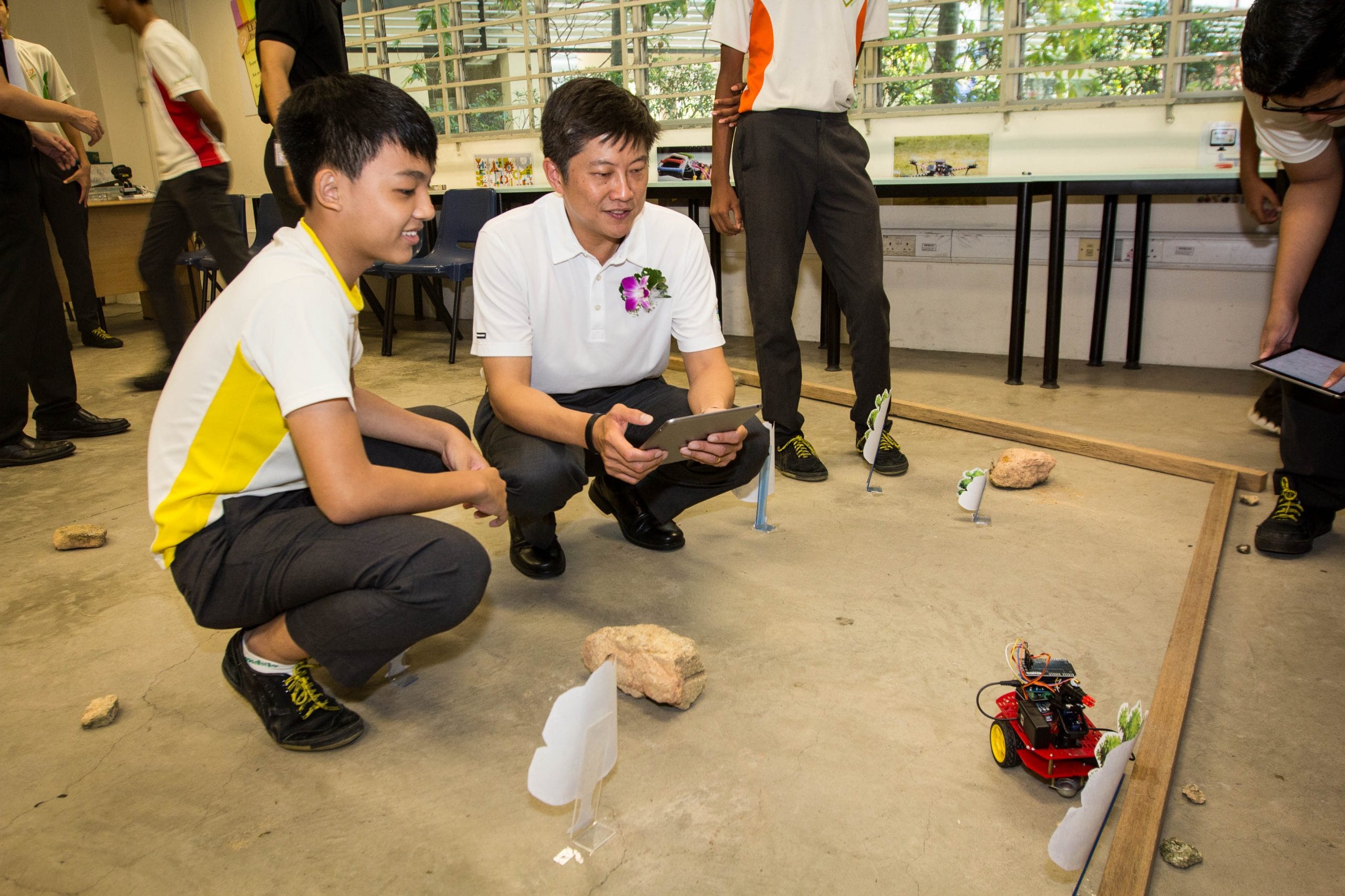 A guest speaker or visitor and a student crouch together watching a small red robot navigate an obstacle course with rocks and markers.
