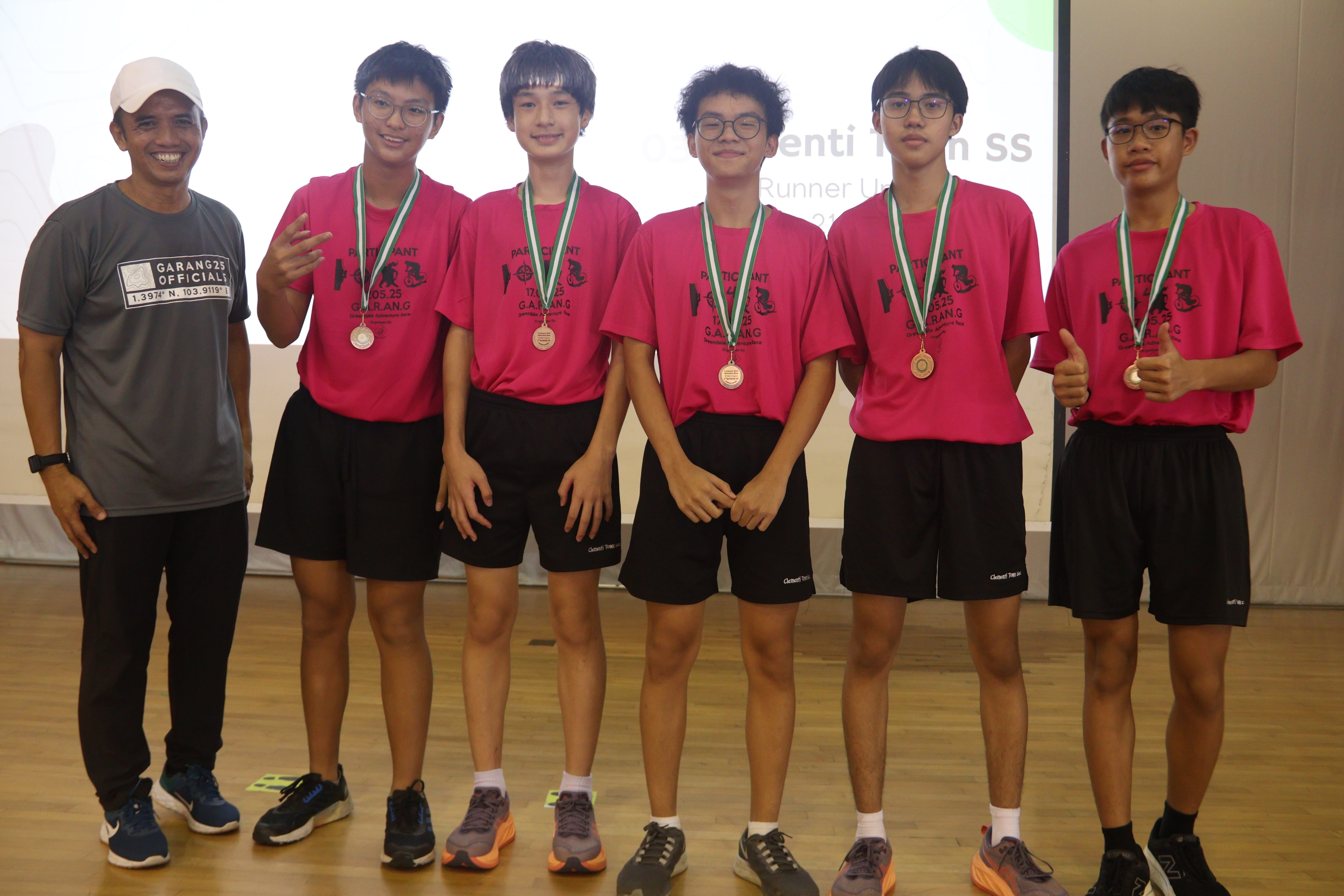 Five smiling students wearing pink t-shirts and medals pose for a group photo alongside an official on a stage.