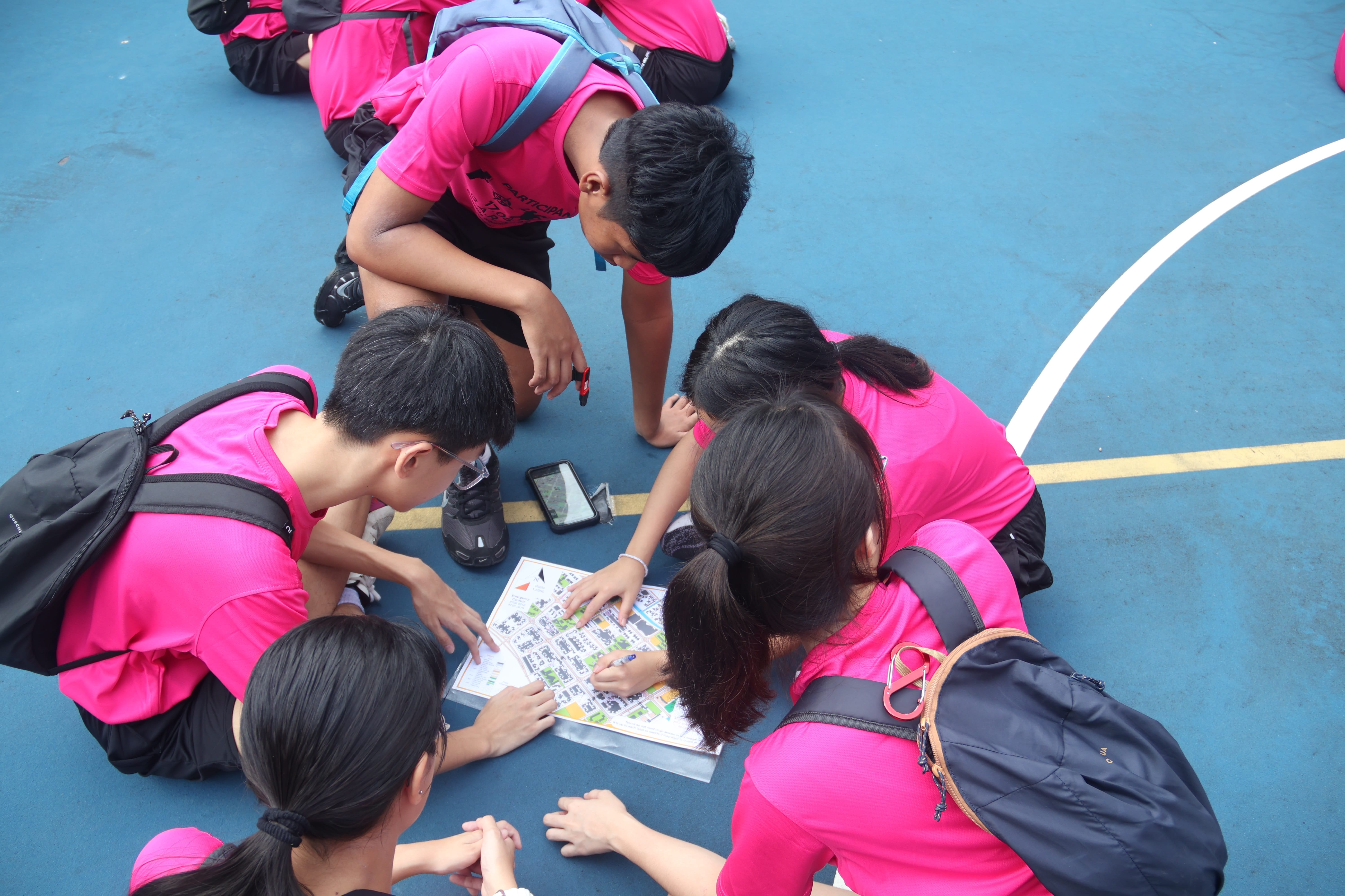 group of students wearing pink t-shirts huddle on a blue outdoor court to read a map and navigate together.