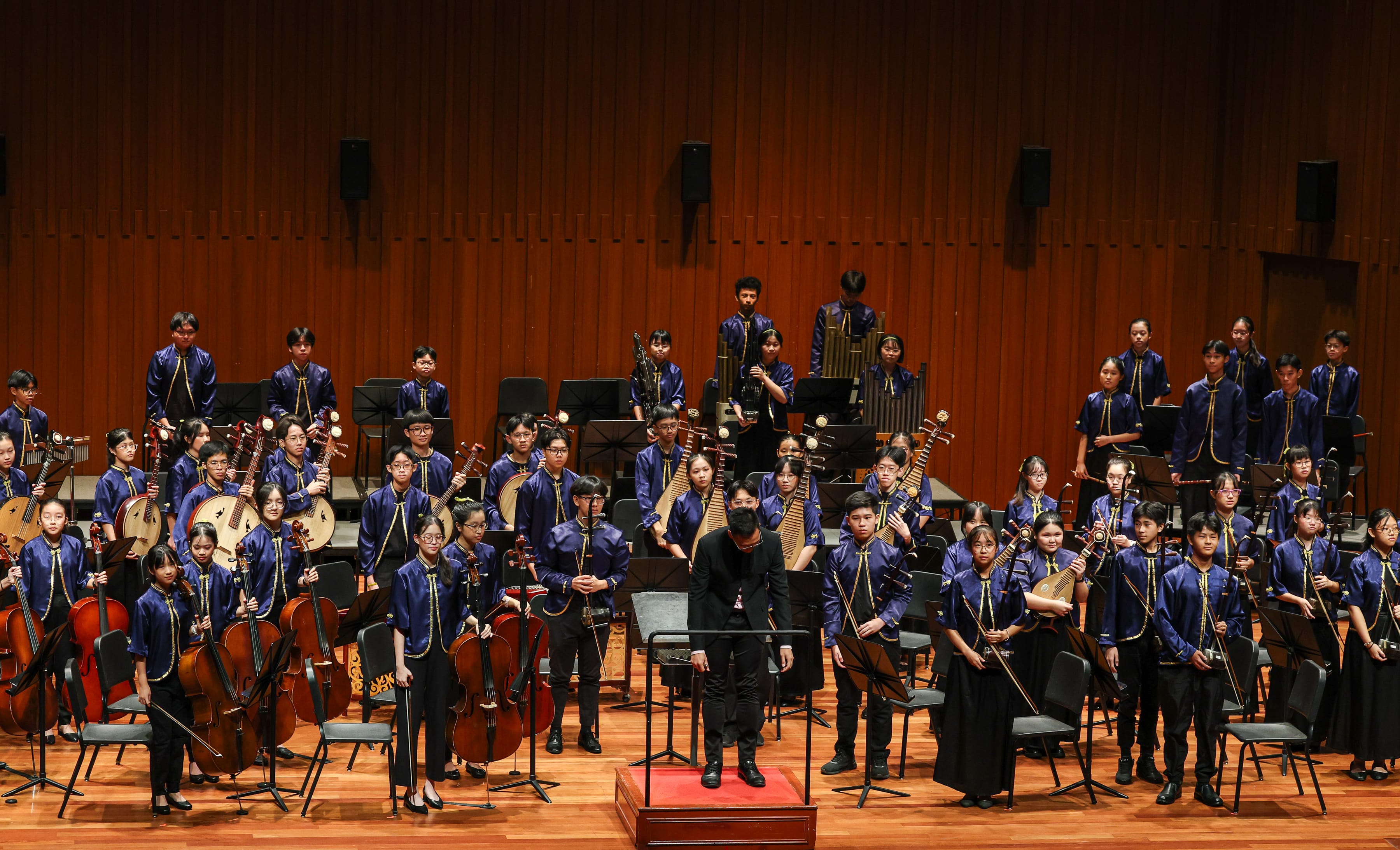 A large youth Chinese orchestra in blue uniforms stands on stage with a conductor bowing at the front.