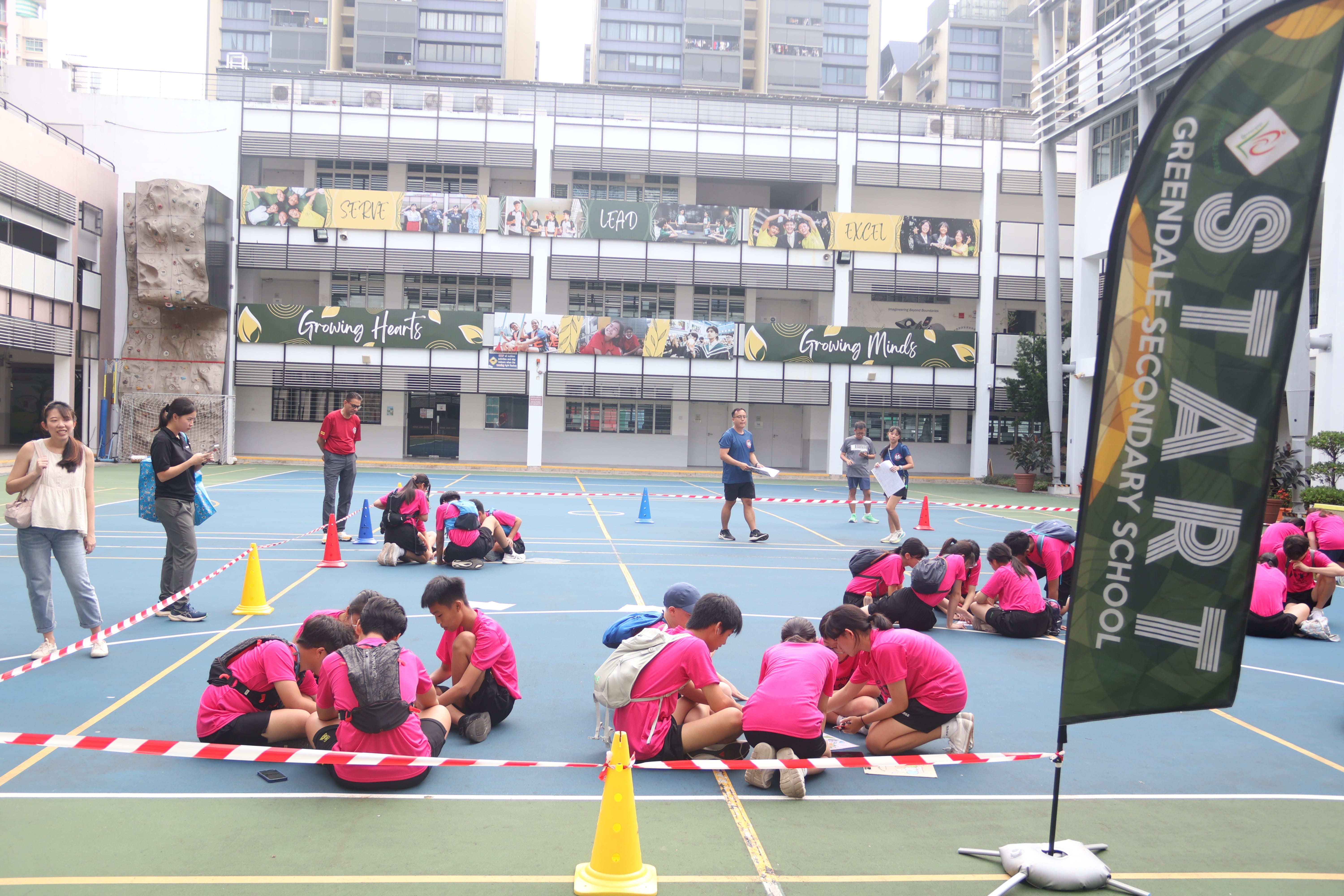 Several groups of students wearing pink t-shirts sit on a blue outdoor court working on activities behind barrier tape, with school buildings and a "START Greendale Secondary School" banner visible.