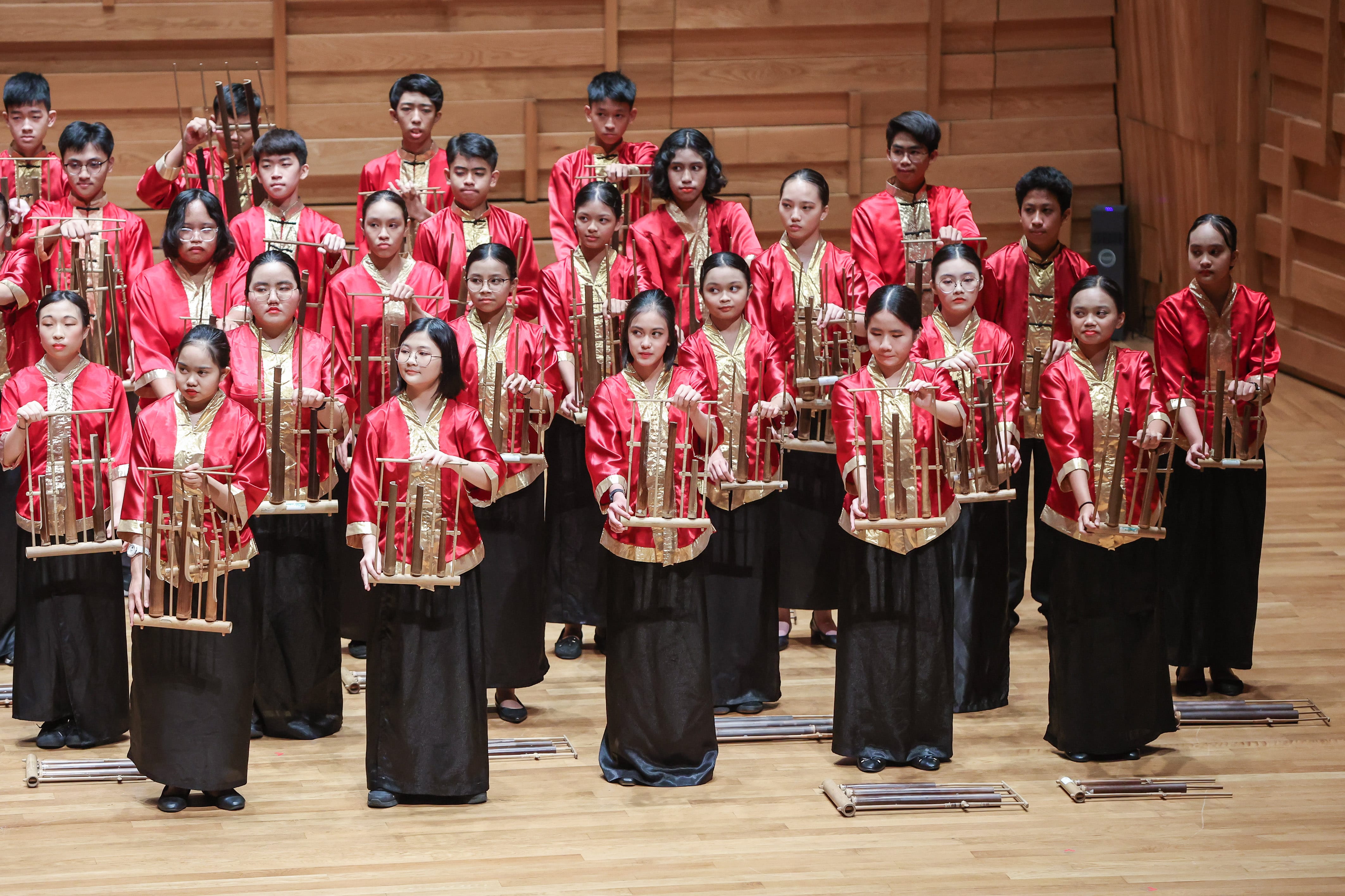 Large group of young performers in red and gold costumes holding bamboo angklung instruments on a wooden stage.