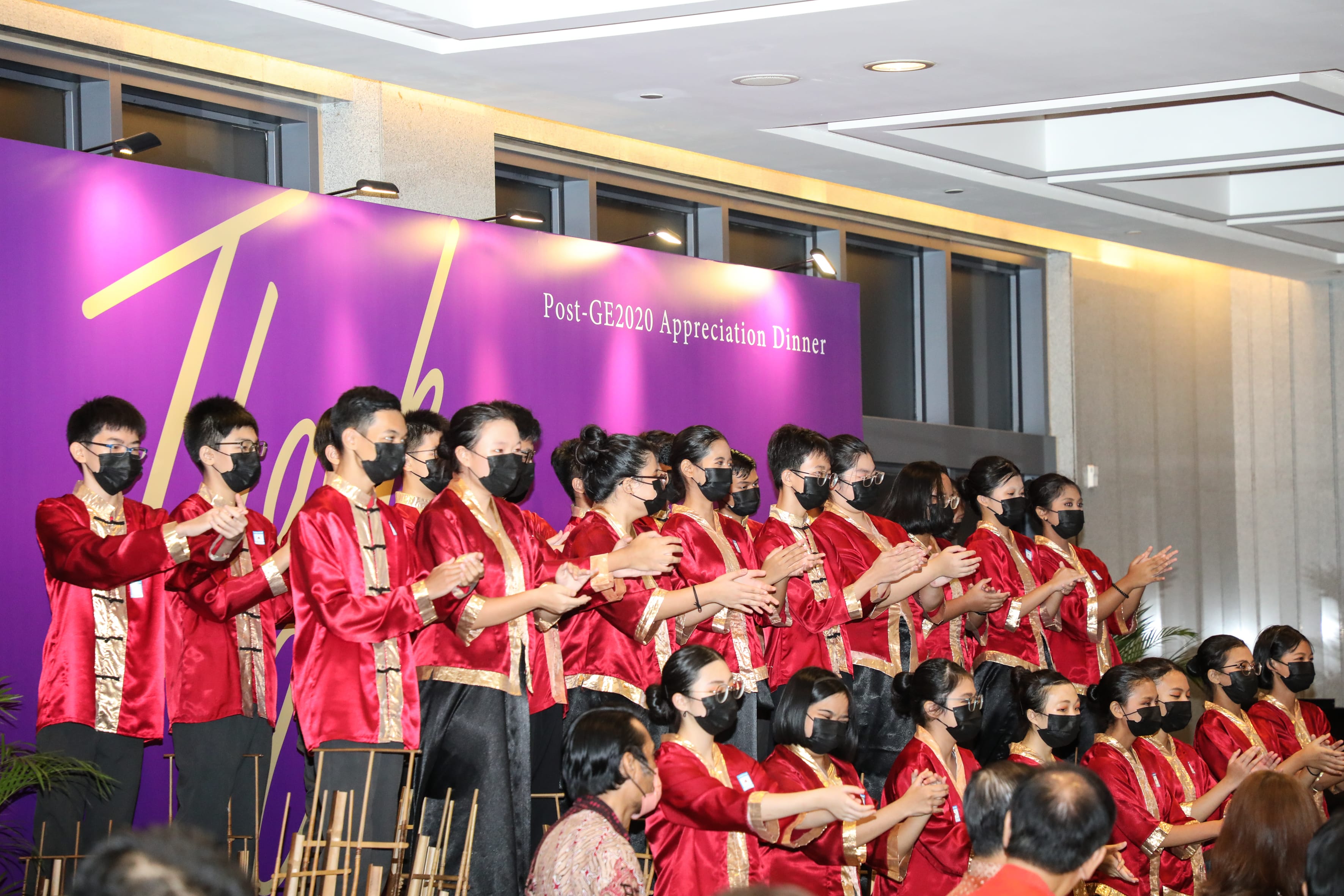 Group in red and gold outfits wearing black masks, clapping on stage before a purple backdrop.