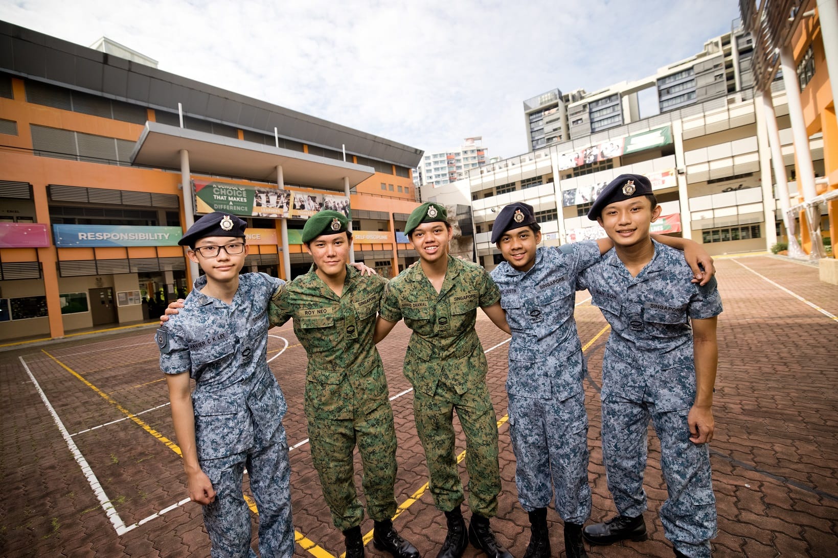 Five young cadets in military uniforms and berets smile together on a school courtyard in Singapore.