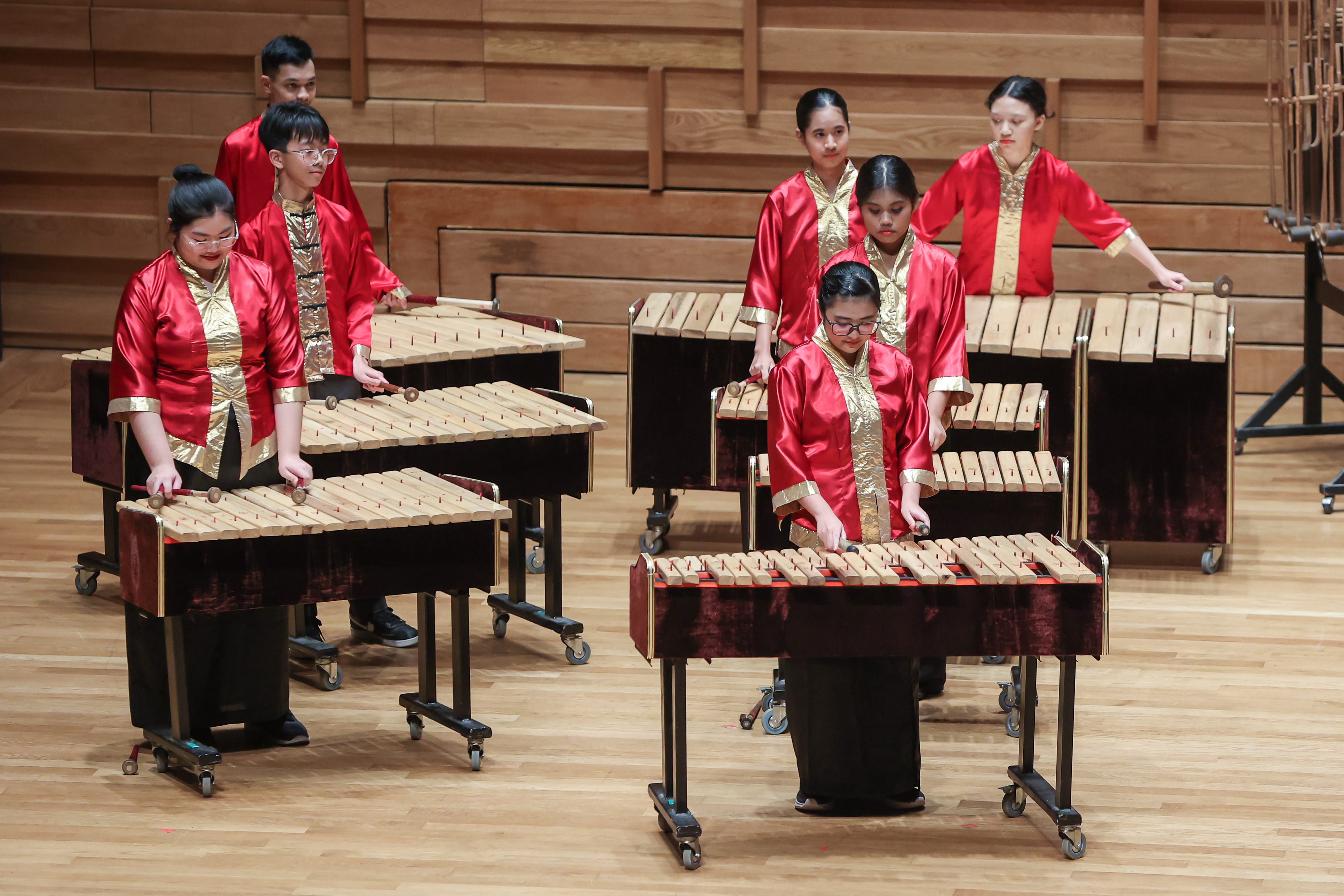 Several young performers in red and gold outfits play marimbas on a wooden concert stage.