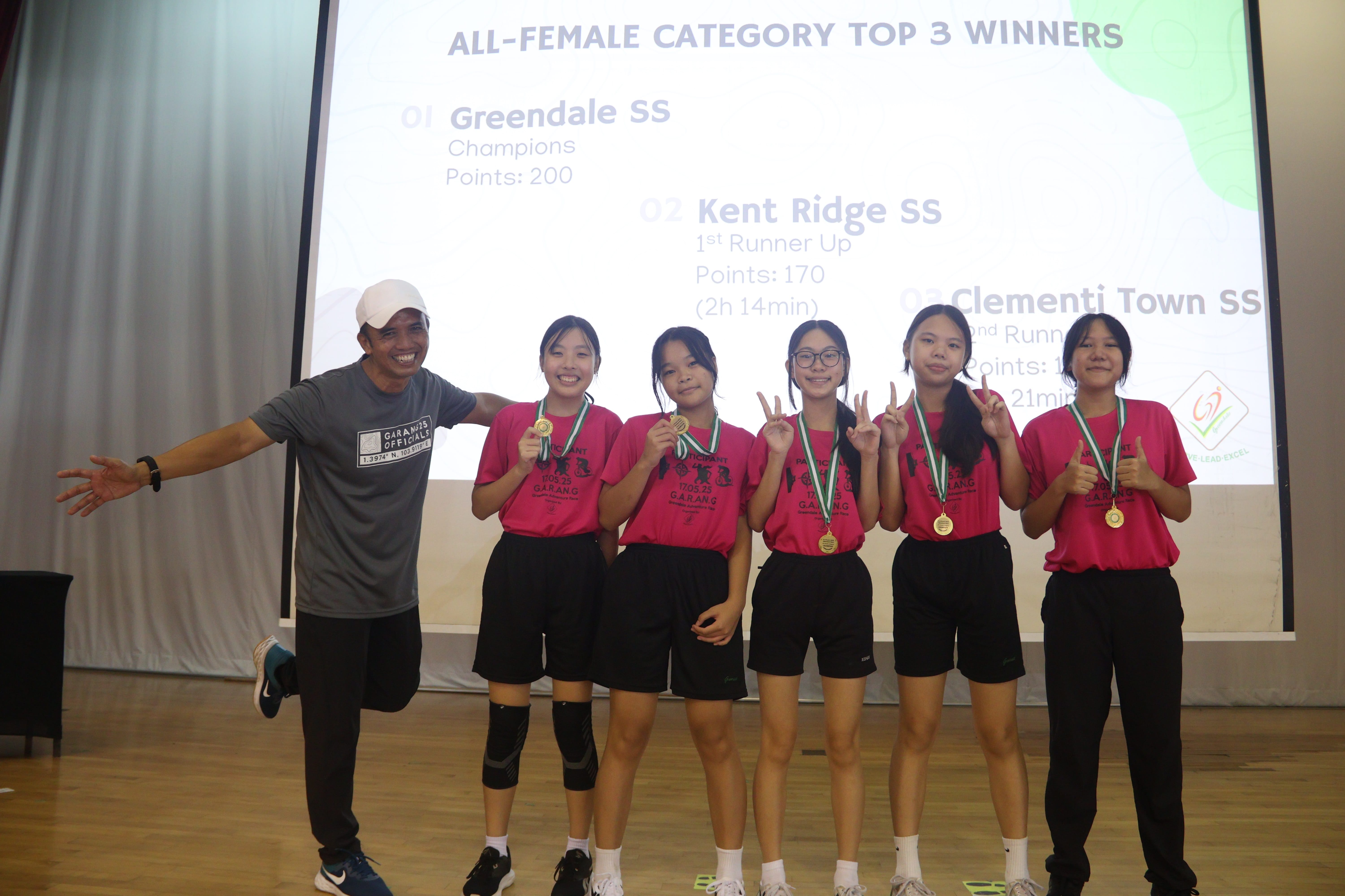 Five female students wearing pink t-shirts and medals smile and gesture on stage next to an official, with a presentation slide showing the "ALL-FEMALE CATEGORY TOP 3 WINNERS" behind them.